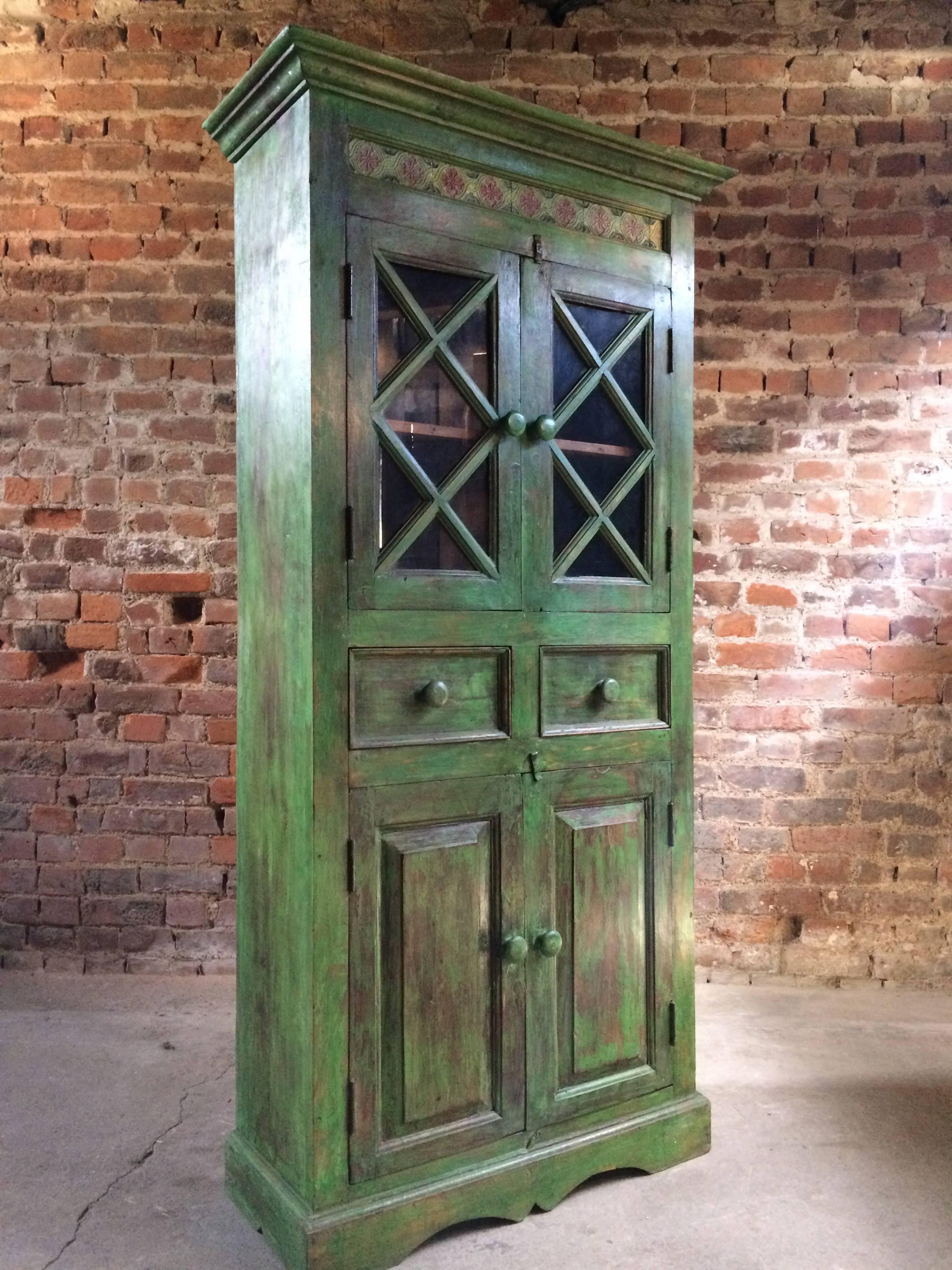 A magnificent early 20th century French painted Teak cabinet, painted in a dark green ground with heavily distressed finish, the corniced top with a tiled decoration over two glazed panelled doors with a single shelf within, over two frieze drawers