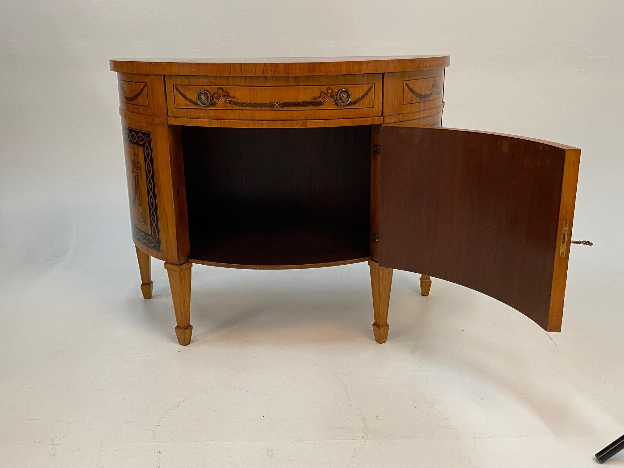 A sublime demilune shaped satinwood sideboard cabinet having magnificent hand painted neoclassical decoration, single drawer and swing out lower door to reveal storage inside.