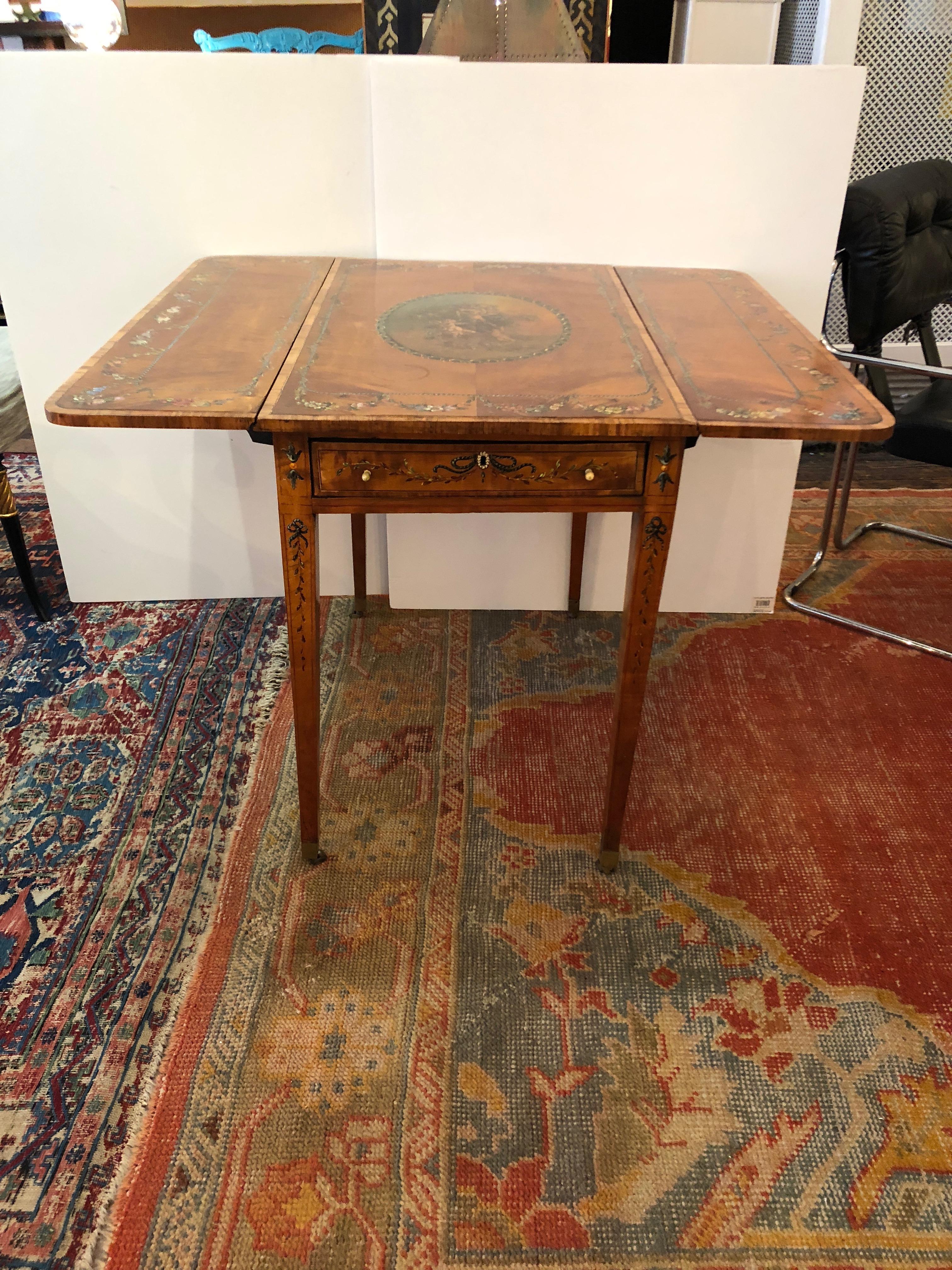 A very beautiful antique French drop-leaf satinwood side table adorned with a central oval landscape having cherubs, while flowers and garlands embellish elsewhere. There is a single drawer with original bone knobs and escucheon, and the tapered