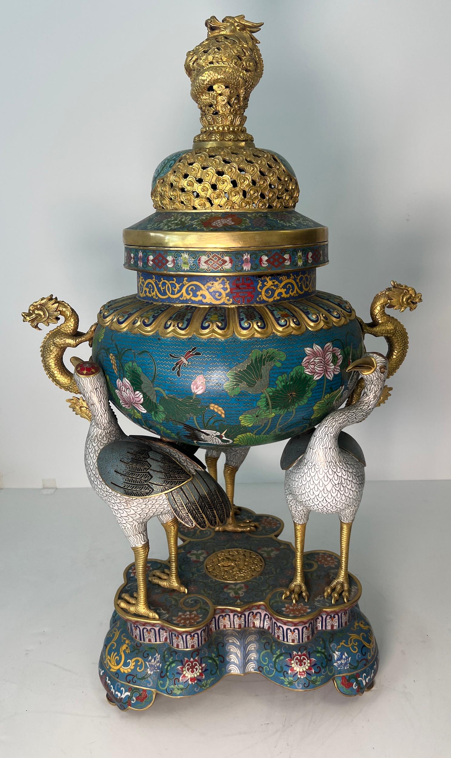 Magnifico incensiere a treppiede in smalto cloisonne e bronzo dorato con coperchio. In stile Qianlong, con un'imponente fusione e sostenuta dalle spalle di tre gru. Il corpo globoso è smaltato a colori con una scena continua di laghetto di loto,