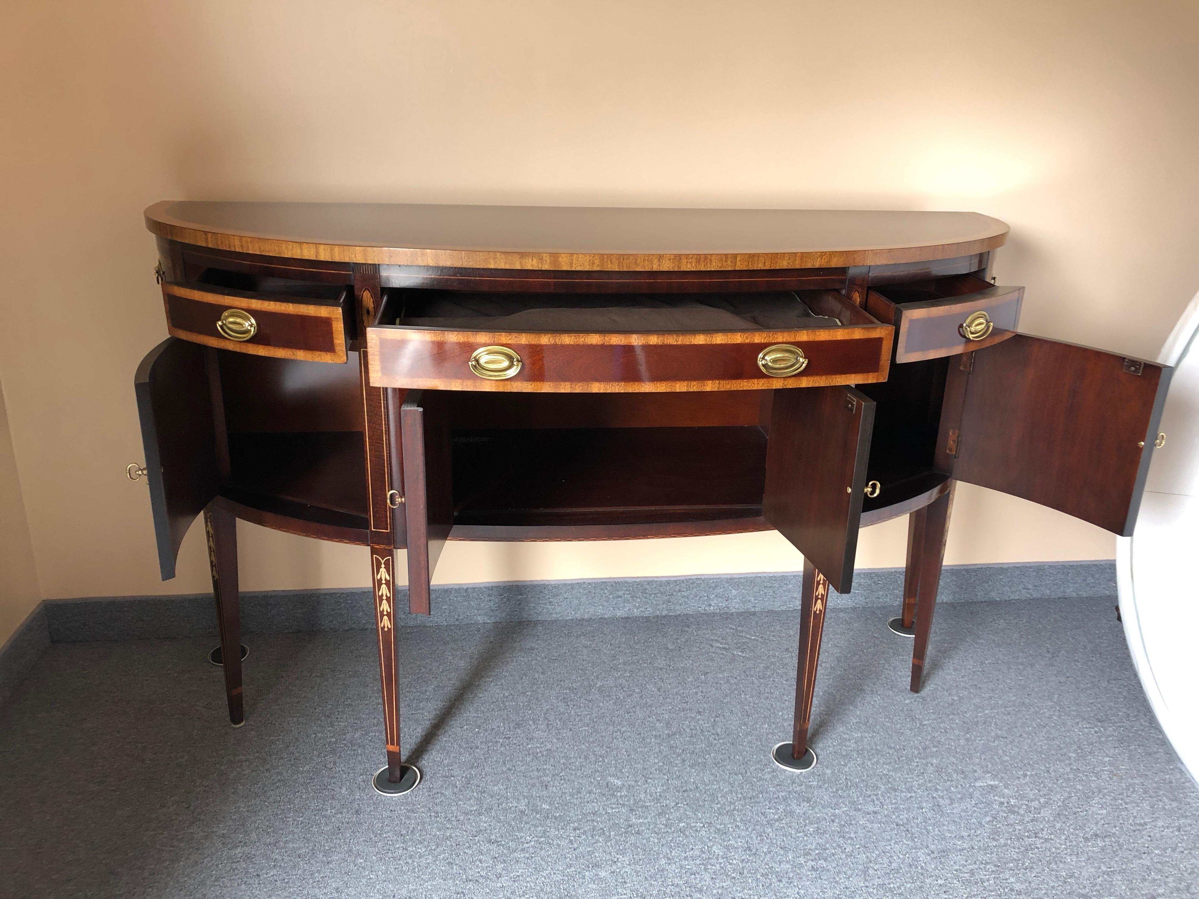 A gorgeous mahogany demilune sideboard having flame mahogany doors, lovely ebony and satinwood borders, highly polished brass hardware and decorative medallions. Three drawers run along the top above 4 doors that open to reveal storage inside. The