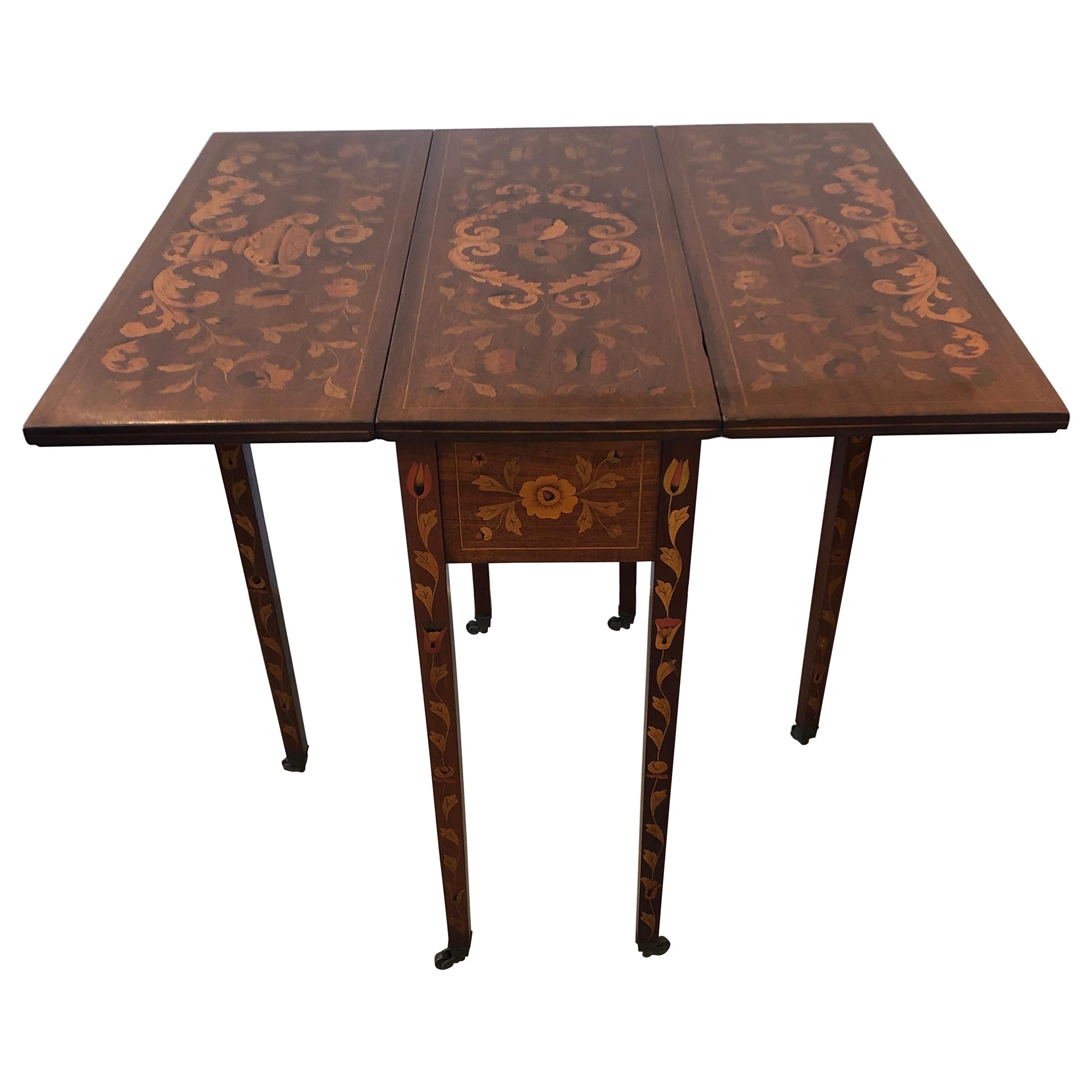 A magnificent 19th century drop-leaf side table from the Netherlands having incredible Dutch marquetry inlay work including leaves, flowers, and urns in mahogany, satinwood, cherry and ebony woods. There is one dovetailed drawer and brass casters on