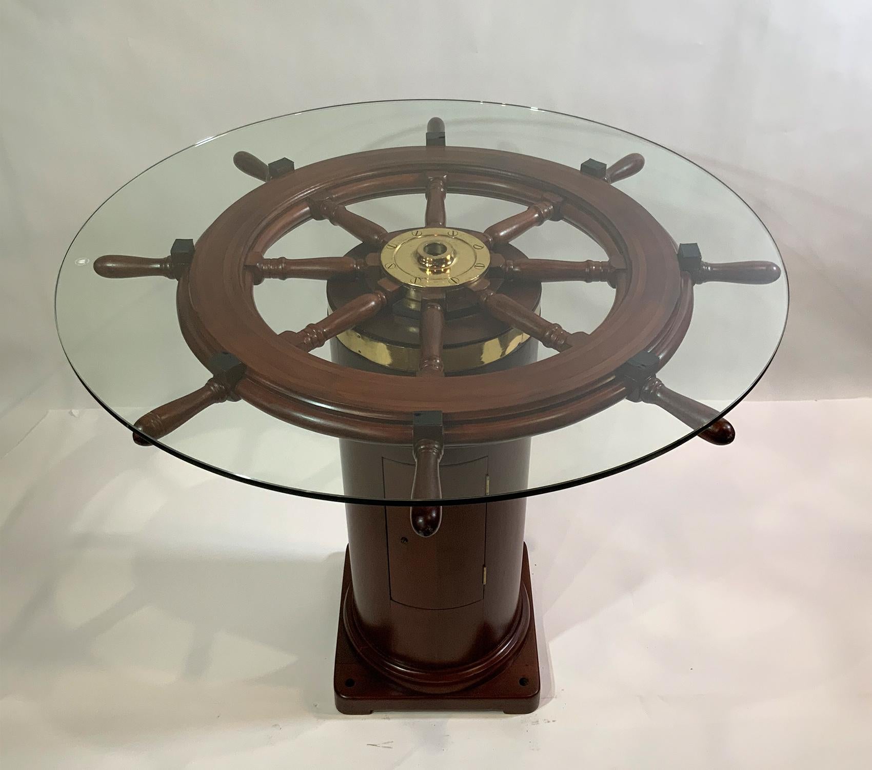 Antique ships wheel fitted to a binnacle base creating a custom bistro table. The wheel and binnacle base have been meticulously sanded, stained and varnished. The brass hub has been meticulously polished and lacquered. A tempered glass top one half