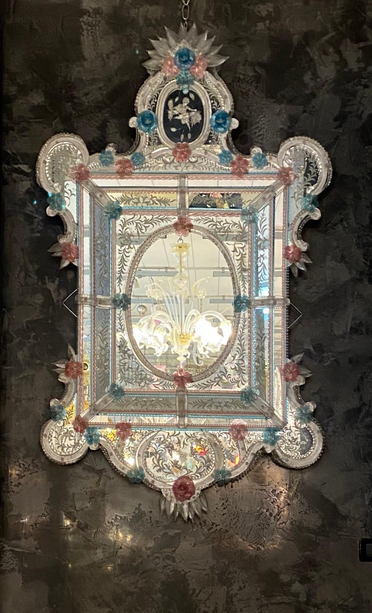 Ce magnifique miroir vénitien présente des motifs floraux gravés qui ornent le cadre du miroir. Le long des bords du cadre se trouvent des accents de corde en verre et de nombreuses fleurs roses et bleues en verre.
Exécuté par le grand maître de