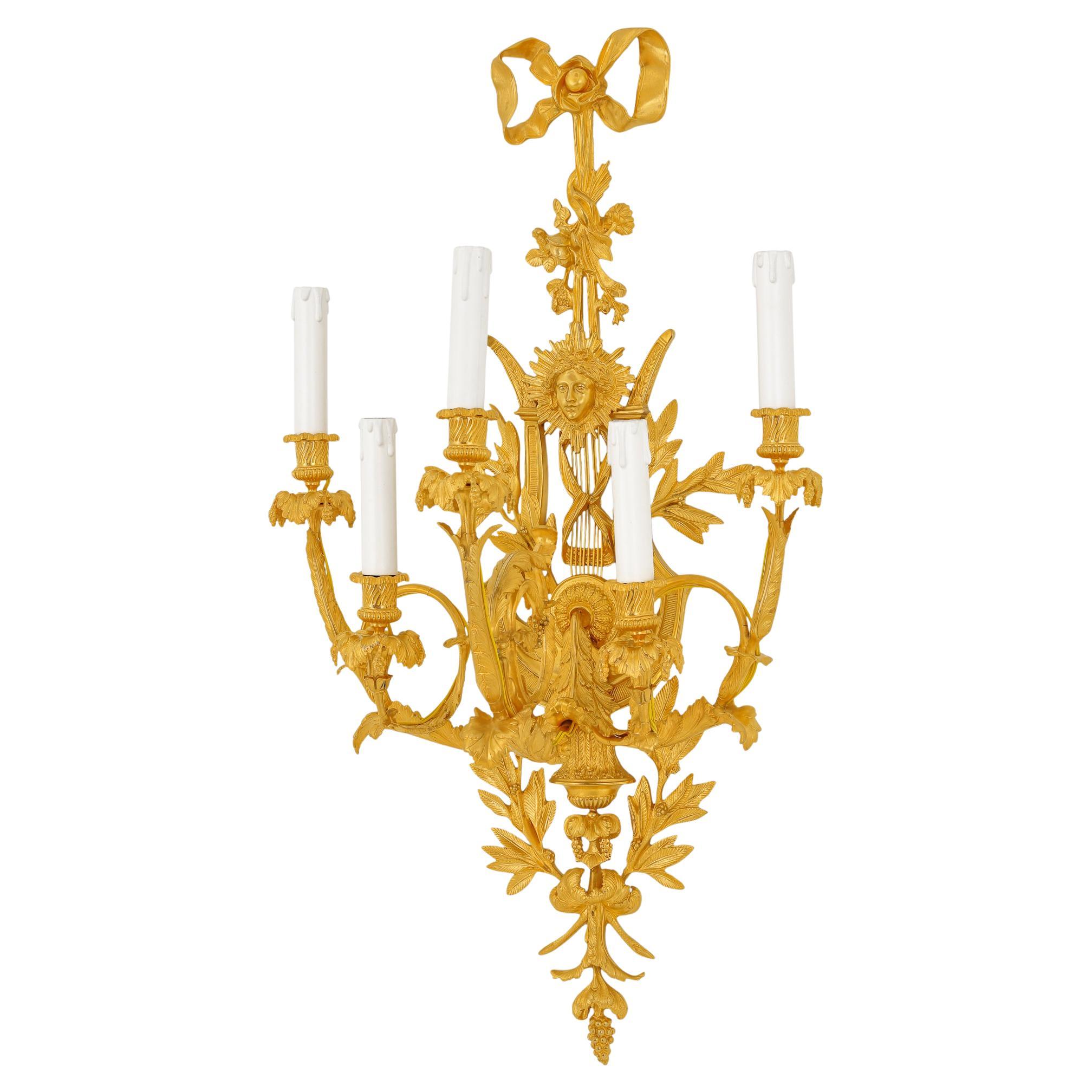 The wall sconce is made of intricately chiseled brass and plated with 24-carat gold, featuring natural elements such as olive branches and the face of King Louis XVI on a shield to convey a sense of grandeur. This wall lamp is a luxurious piece,