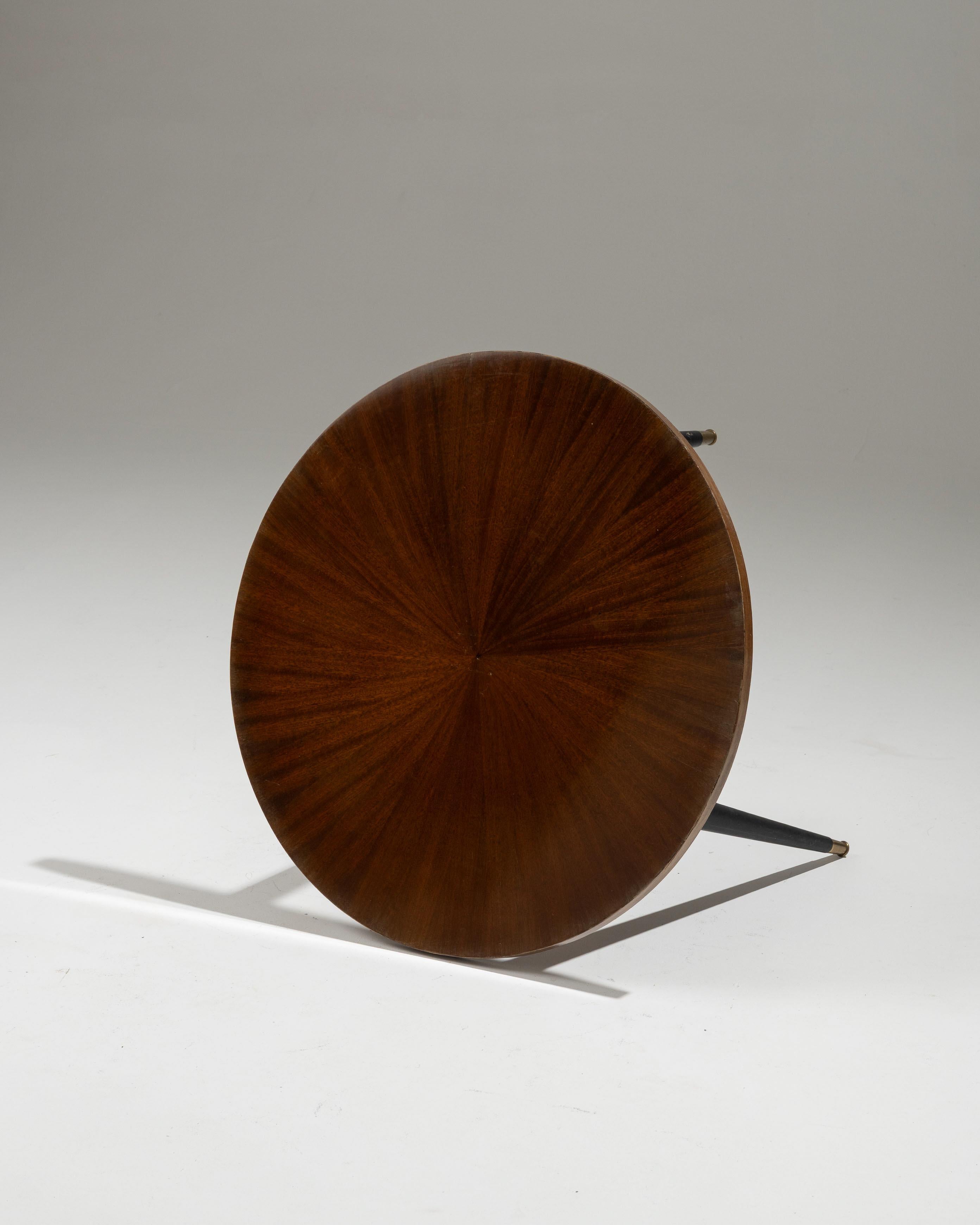 Table basse tripode en placage d'acajou et bronze, années 1950. Le plateau circulaire en acajou est décoré d'une frise rayonnante et repose sur trois pieds fuselés en bronze laqué noir, vissés dans le plateau. Une pièce élégante qui joue sur les