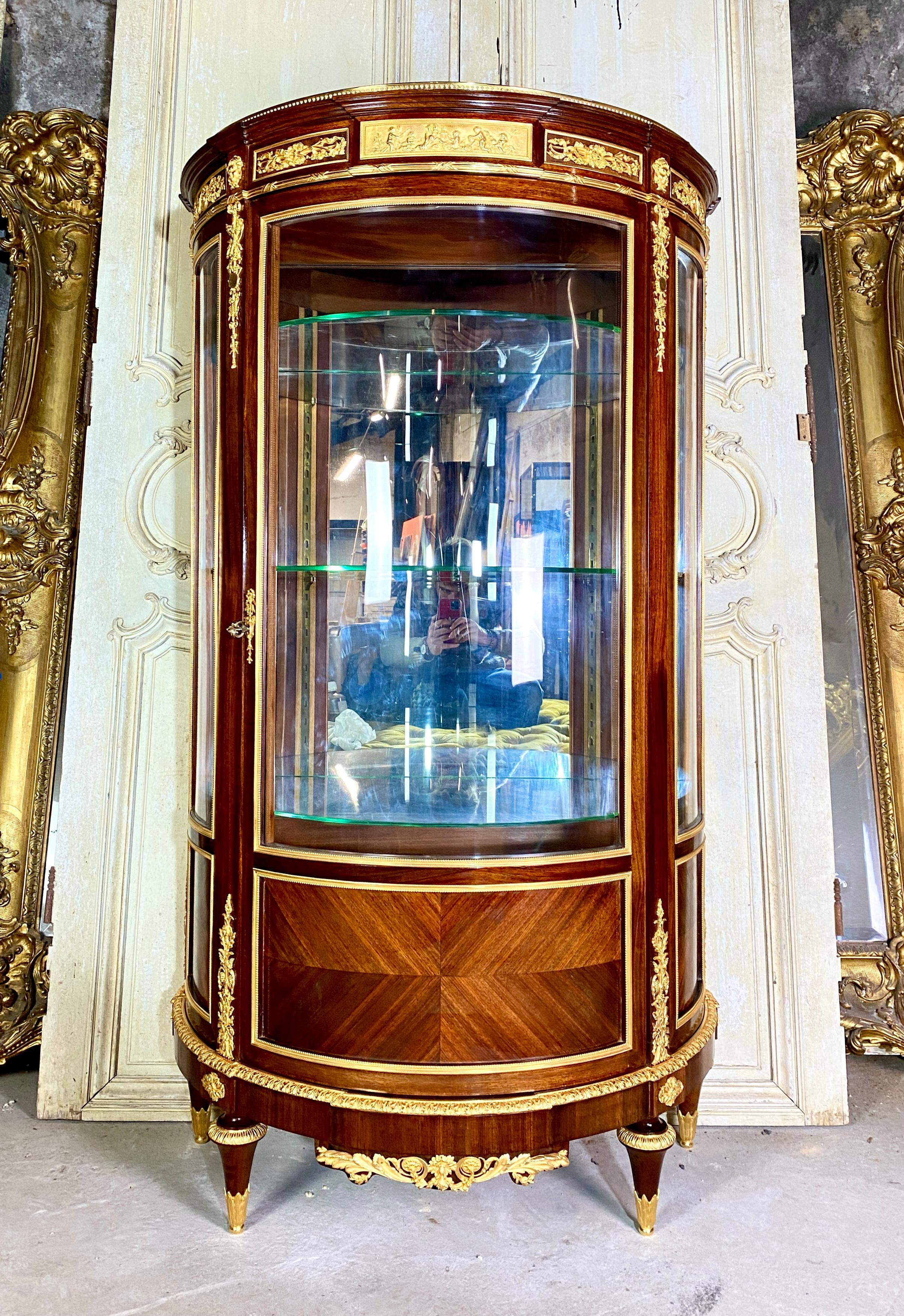 Mahogany half-moon-shaped showcase adorned with many finely chiseled and gilded bronzes with a bas-relief in the center representing a scene with puttis in the style of Clodion. It is made up of three curved panes and opens with a central door. It