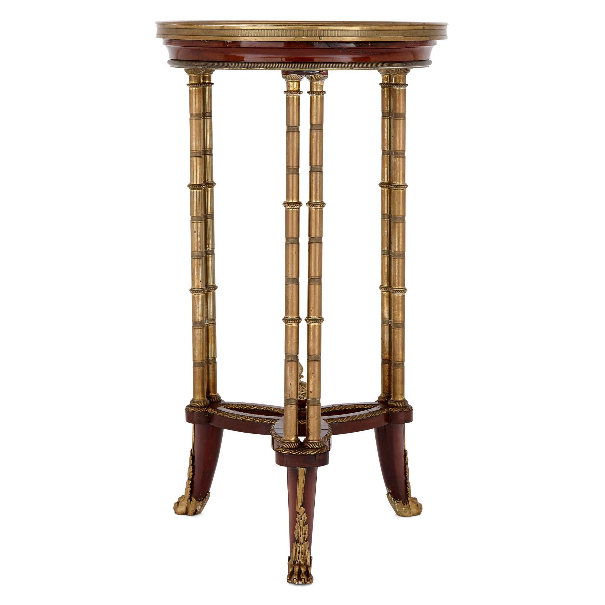 This circular side table is surmounted by an inset onyx top, which is bounded by a gilt bronze ring and is set above a mahogany apron. The table stands on three two-column gilt bronze legs, which in turn meet the ground on gilt bronze hoof feet. The