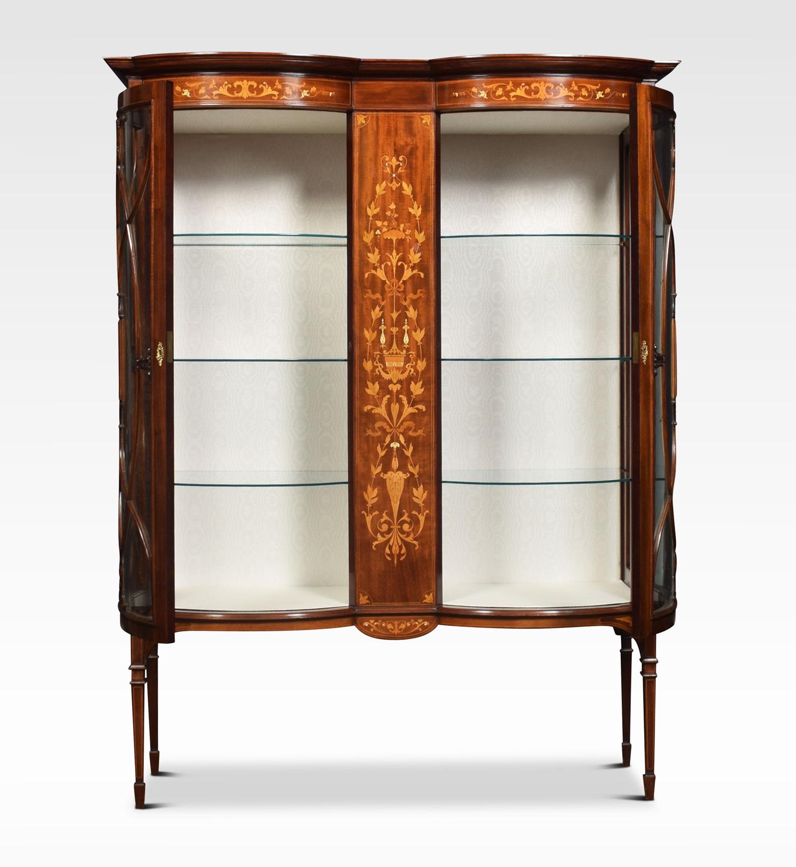 Mahogany display cabinet, having a double bow-fronted shaped top over central floral marquetry panel. Flanked by bowed glass doors enclosing three glass shelves to each side and watermark silk upholstery. All raised up on tapering legs terminating