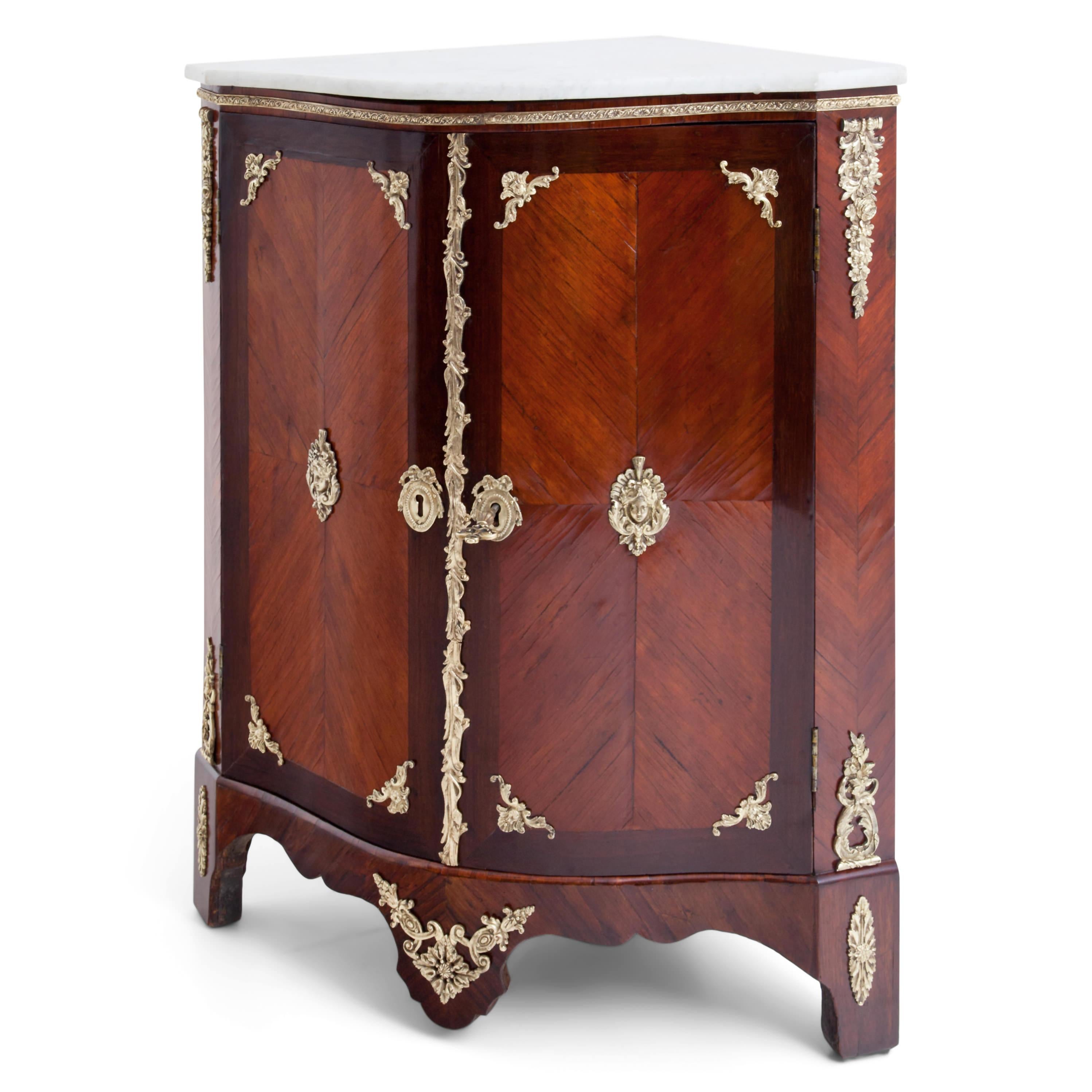 Two-door corner cupboard on wavy cut-out apron with marble-top and mahogany veneered, rounded body. Appliqués of fire-gilded bronze in the form of tendrils, rosettes and mascarons. The marble top is recent of the 19th century. Under the plate