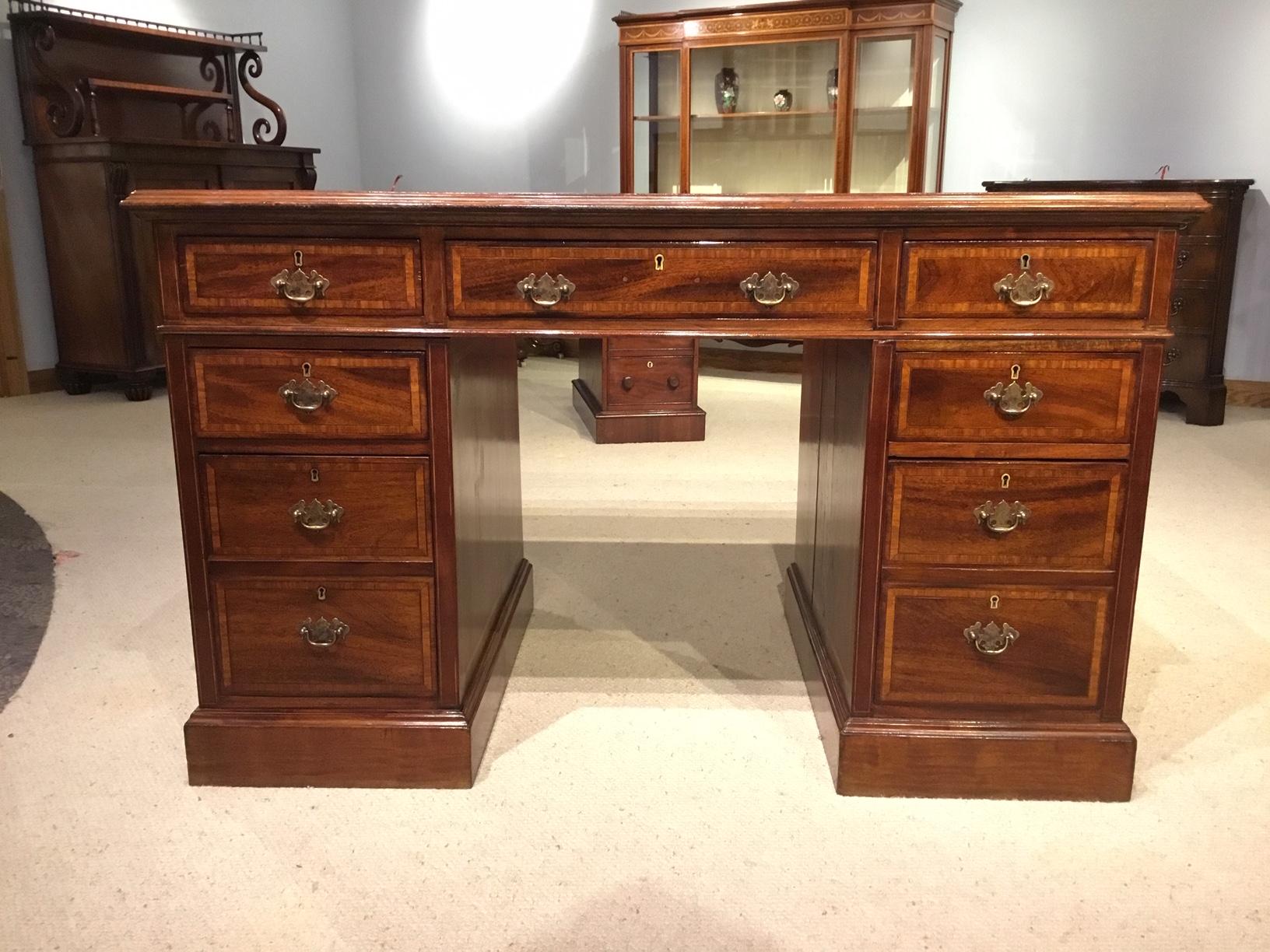 A mahogany and satinwood banded Edwardian period antique pedestal desk. Having a rectangular solid mahogany top cross banded in satinwood and with an inset green leather writing surface with gilt tooled detail. With an arrangement of nine