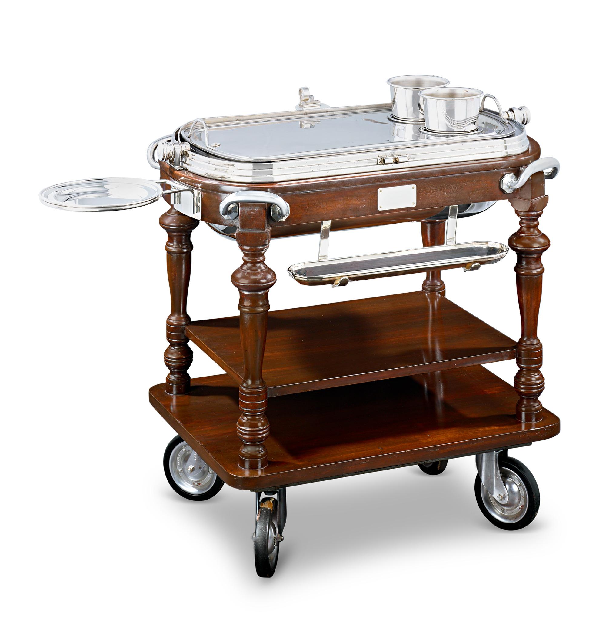 This exquisite mahogany and silverplate carving trolley unveils a generously sized serving space when opened. Resting on an impressive mahogany stand, it features an au jus plate, detachable serving pots and three silver-plated kerosene burners. A