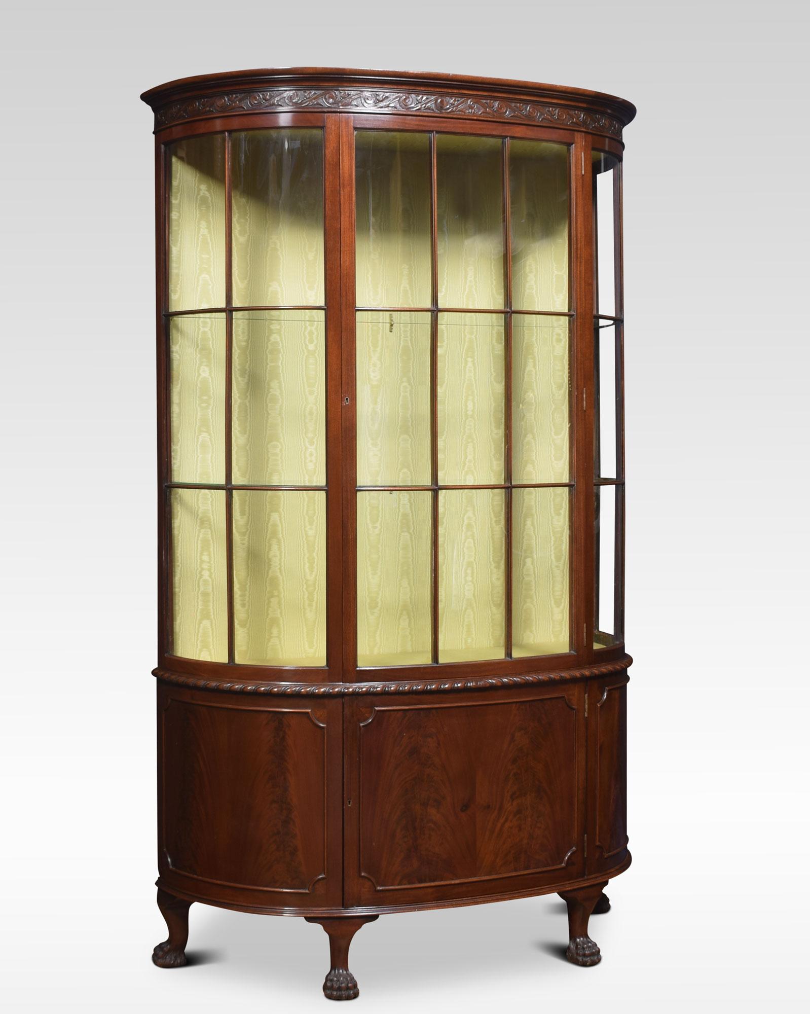 Early 20th century mahogany bow fronted display cabinet, having moulded cornice, above large glazed door opening to reveal watermark silk upholstered interior and two glazed shelves. The base section fitted with a central cupboard door. All raised