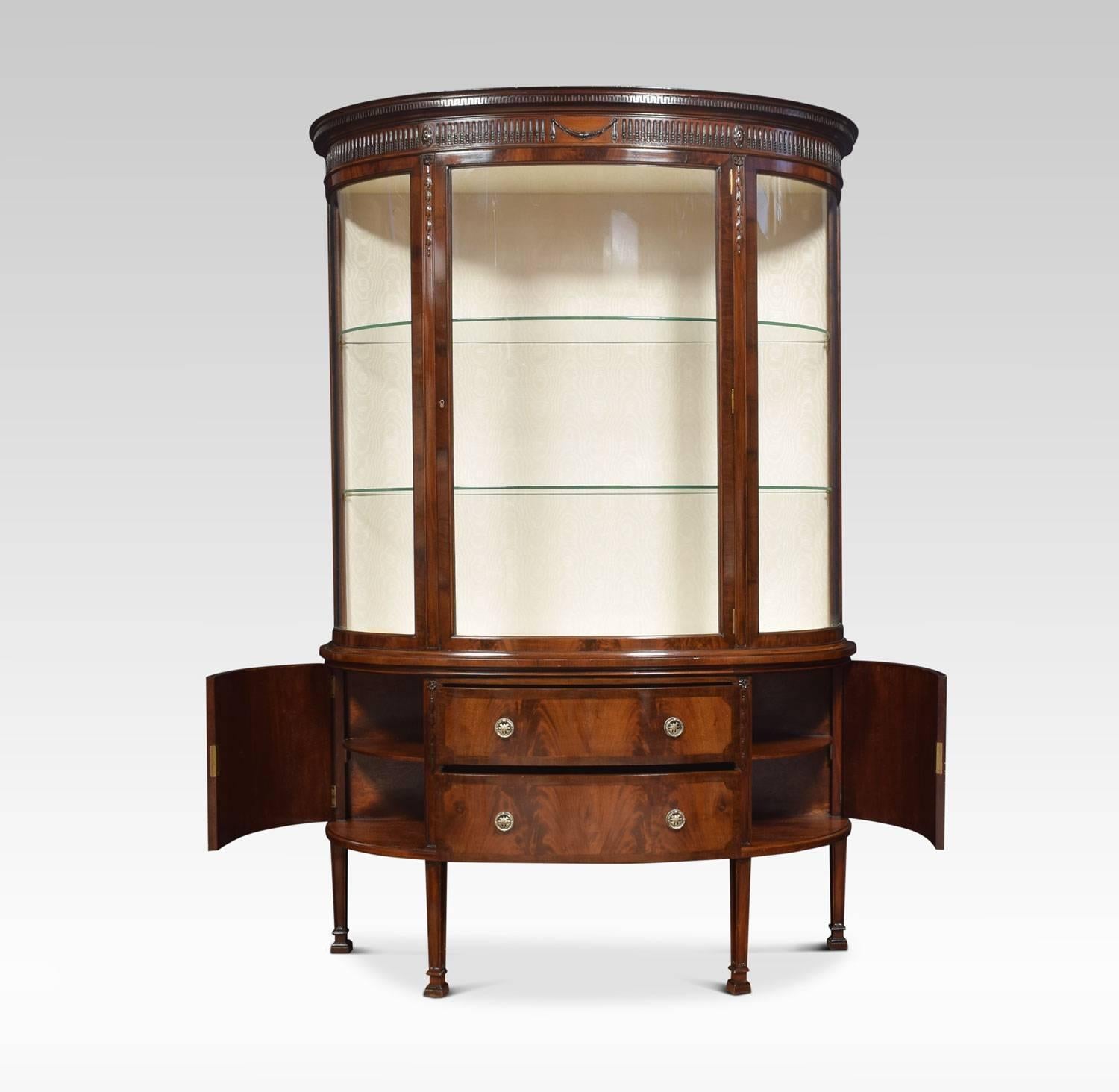 Mahogany bow fronted display cabinet the moulded cornice with central urn and floral swag carving above large central single door opening to reveal two glazed shelves and upholstered interior. The base section having two central draws with tooled