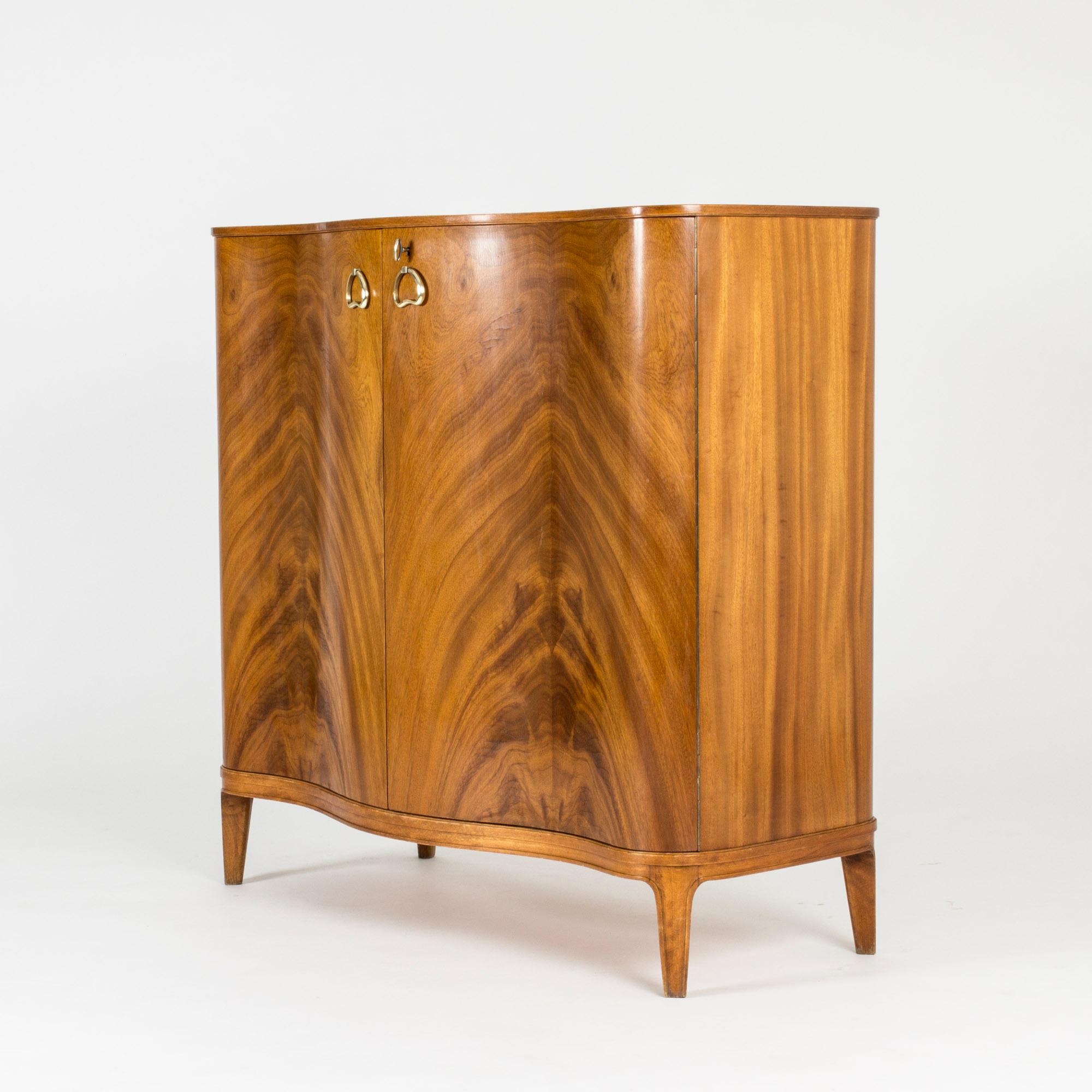 Beautiful mahogany cabinet by Axel Larsson, with dramatic veneer on the door fronts and lovely brass handles. Bulging shape and shelves that follow the same shape inside. Luxurious interior with compartments for small objects, like cufflinks, and