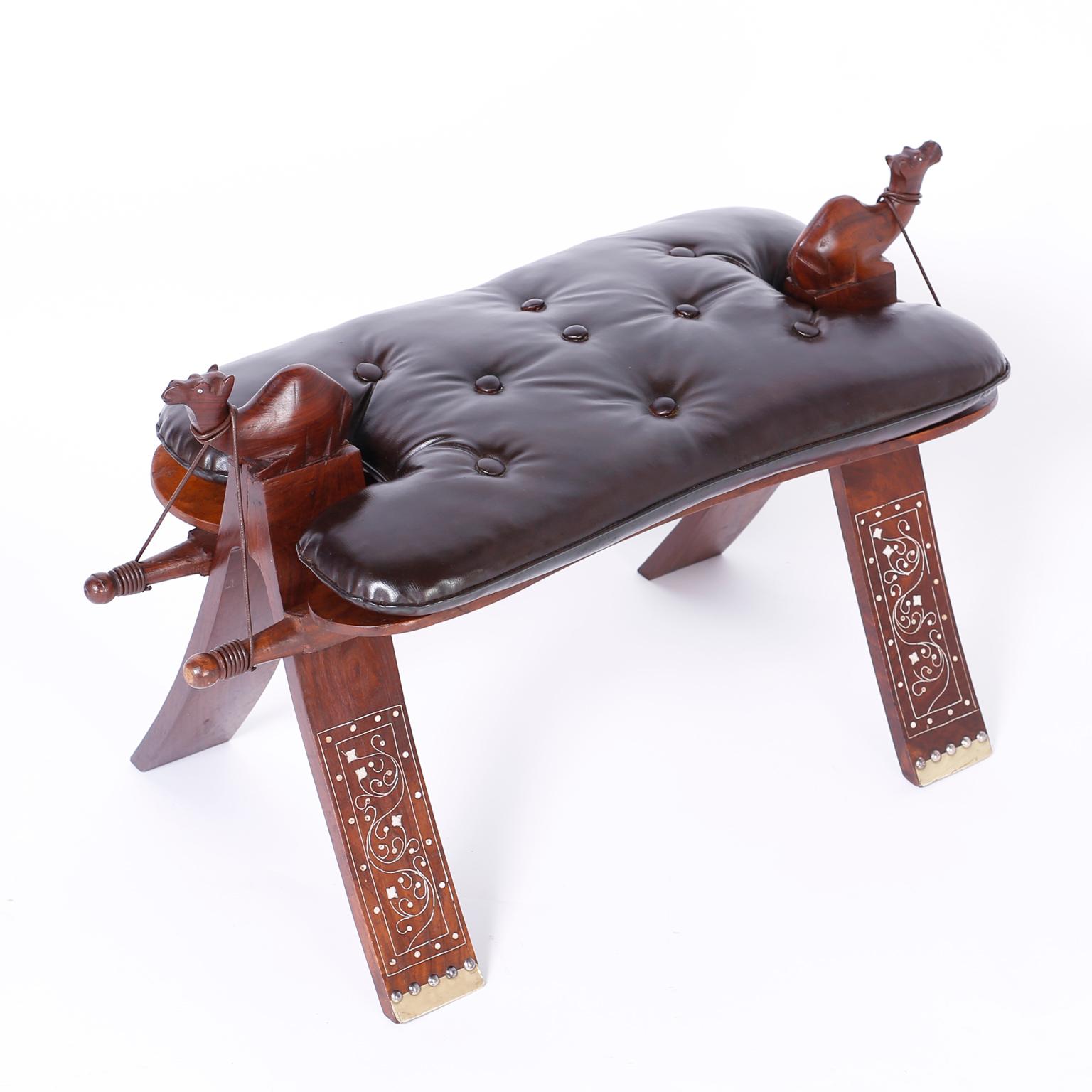 Camel saddle foot stool crafted in mahogany with two carved camels at the top, dark chocolate brown leather button tufted cushion, and splayed legs decorated with floral brass inlays.