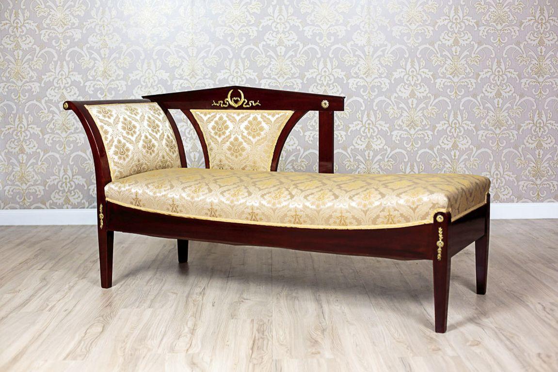 Eine Chaiselongue aus Mahagoni und Weichholz mit Mahagoni-Maserung.
Der Sitz ist gefedert und mit einem beige-goldenen Stoff gepolstert.
Die Form des Möbels ist einfach und bezieht sich auf den Klassizismus.
Die Vorderseiten der Beine und die