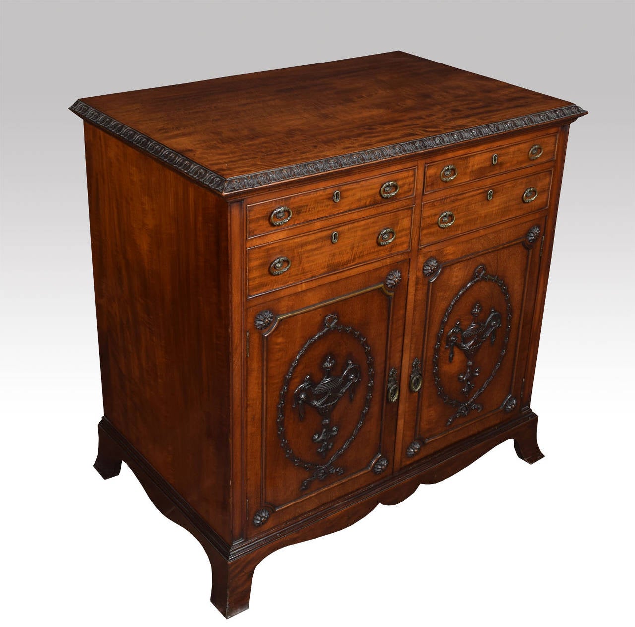 Mahogany commode in the influenced of Robert Adam, the rectangular top having carved edge above four short drawers with tooled brass handles over two doors with classical urns, ribbon and swags mouldings, opening to reveal single fixed shelf Raised