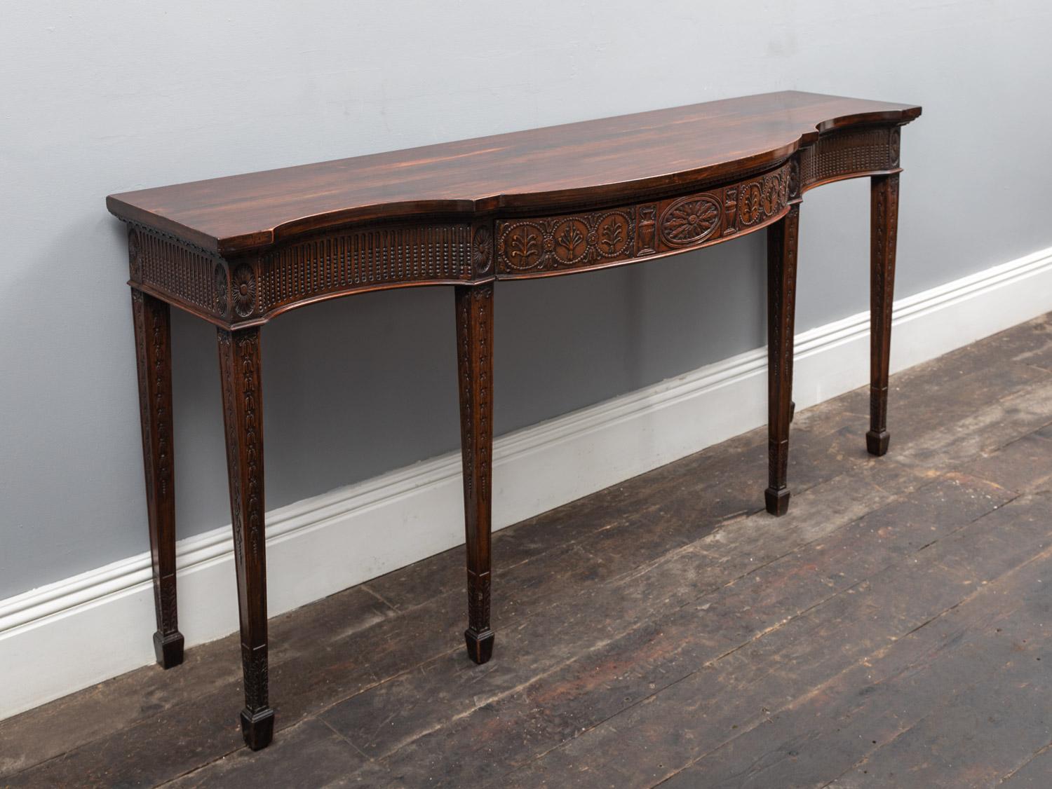 A George III period mahogany side table in the neo-classical Adam style. The moulded serpentine top raised over a carved frieze of classical decoration, the long central drawer finely carved with anthemion's, palmettes and Grecian urns.  The frieze