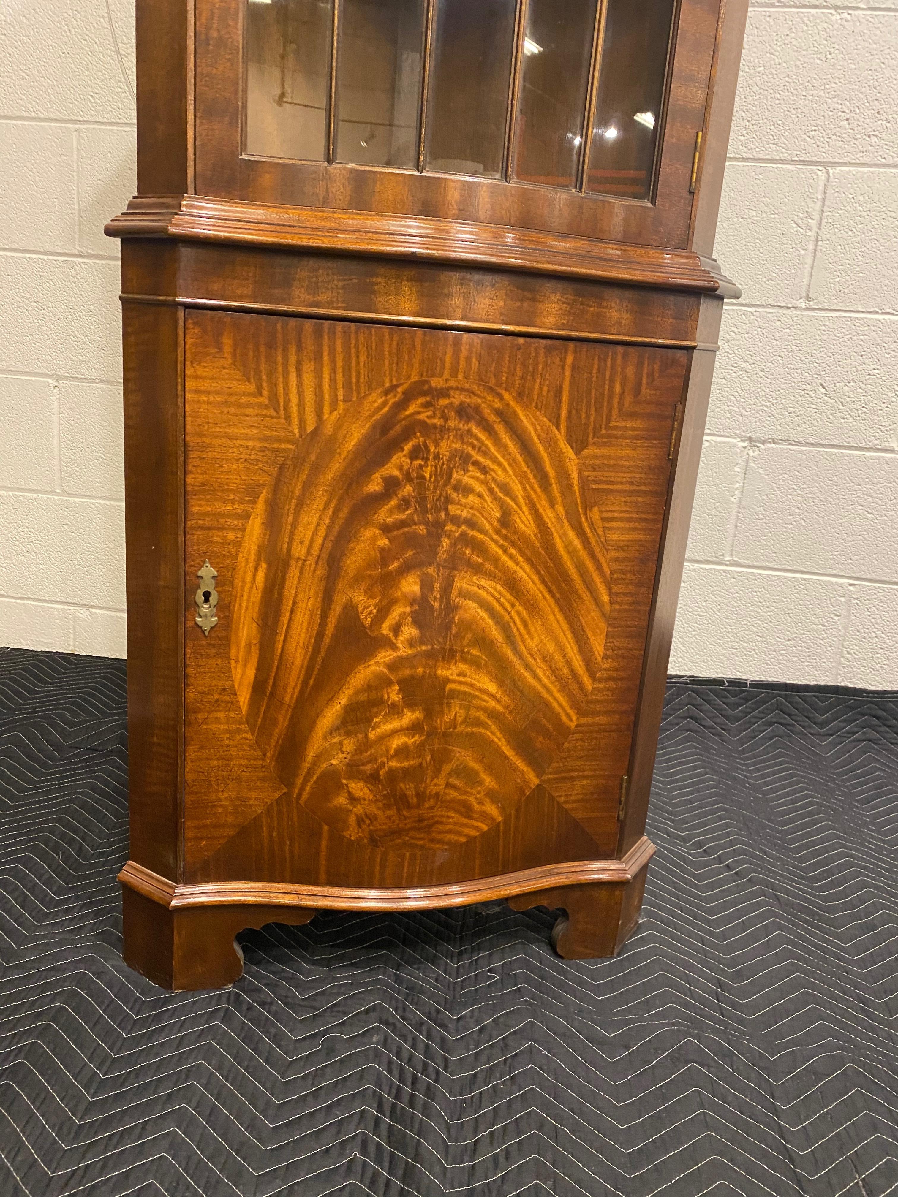 A lovely mahogany corner cabinet with a serpentine shaped curved front face, glass door with individual hand glazed panes. Single door top and bottom, bottom door is a figured mahogany centre panel with a mahogany cross band detail. Dentil moulding