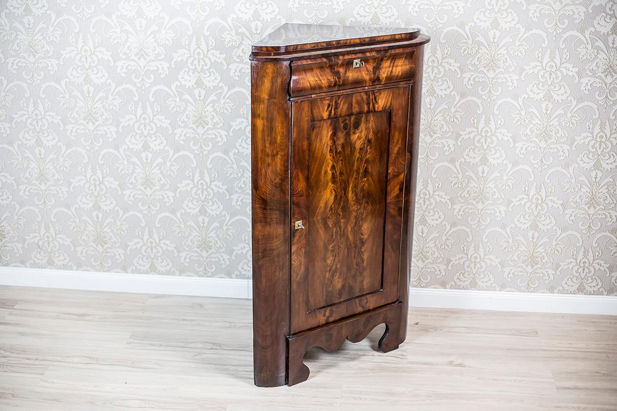 We present you a corner cabinet made of softwood veneered with pyramidal mahogany, dated 1860.
This piece of furniture is of a simple form, one-door, with a drawer under the top with a concave-convex front and rounded corners, and a base with a