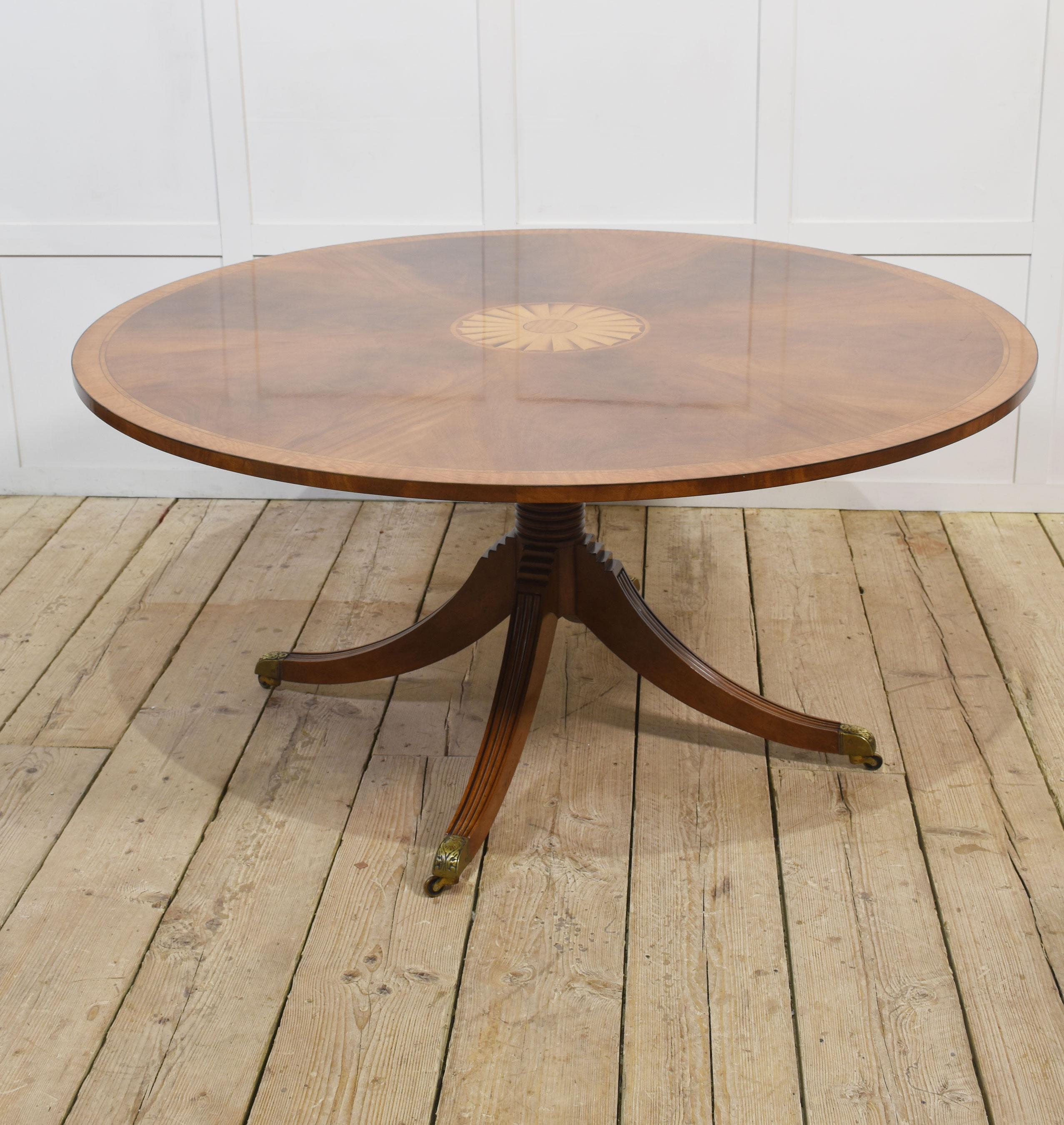 Mesa de comedor circular de caoba con tablero bien tallado e incrustación central rodeada de un borde cruzado. Se eleva sobre una columna torneada con soportes abocinados que terminan en casquillos y ruedas de latón. La mesa tiene capacidad para 6-8