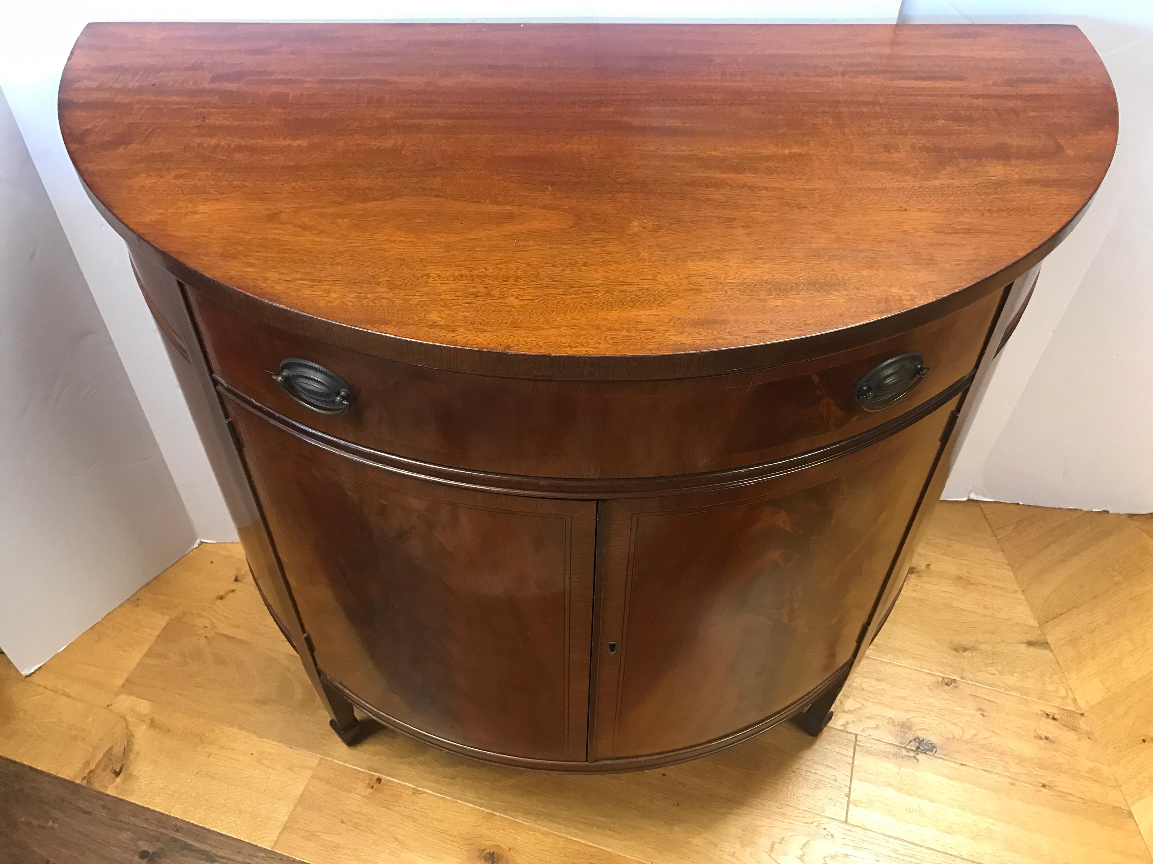 Flame mahogany demilune liquor cabinet features ebonized banding and satinwood inlay. It has one top drawer and two front doors that open to two shelves. Has working lock and key. Made by famed Johnson Furniture Company and hallmarked as such.