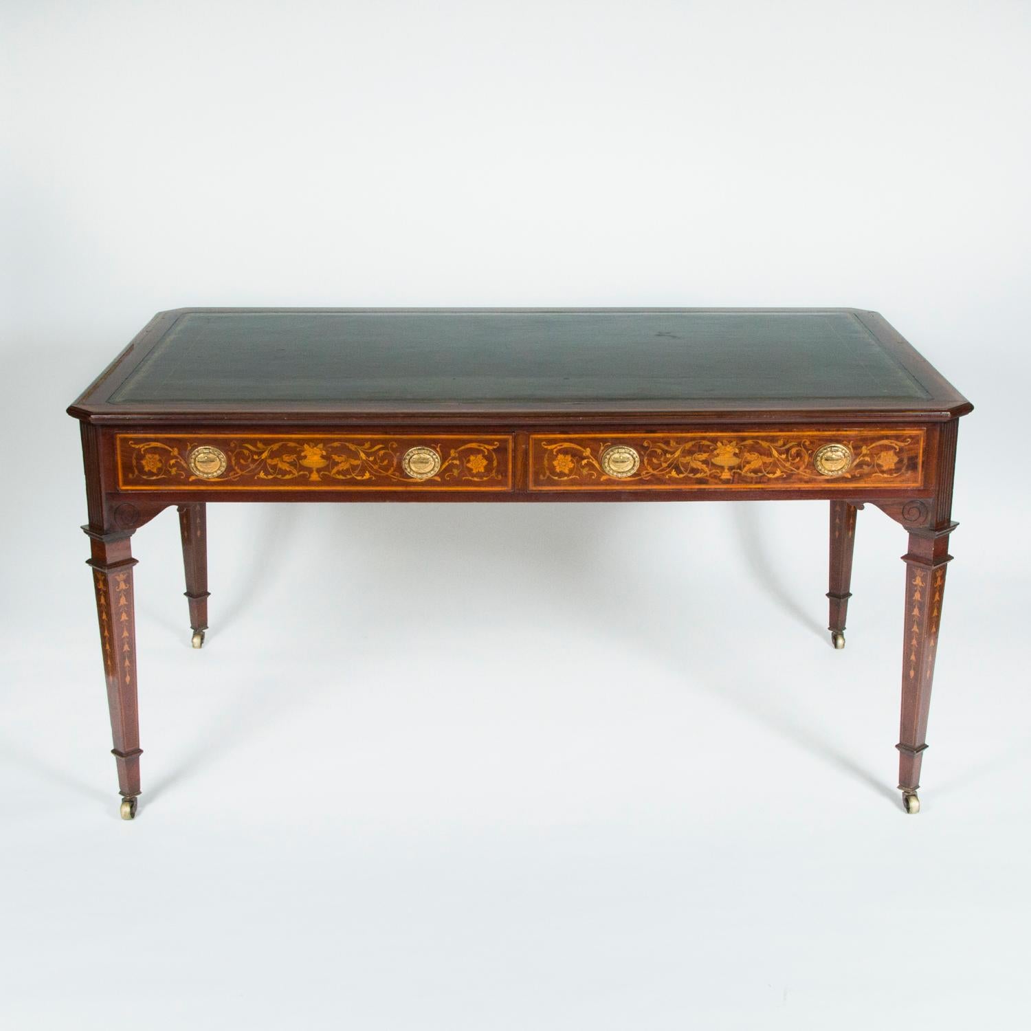 A fine quality freestanding mahogany desk, with detailed floral marquetry and carved decoration the frieze and legs, a green tooled leather top with satinwood banding, and brass castors. 

The frieze conceals two drawers with oval gilt brass