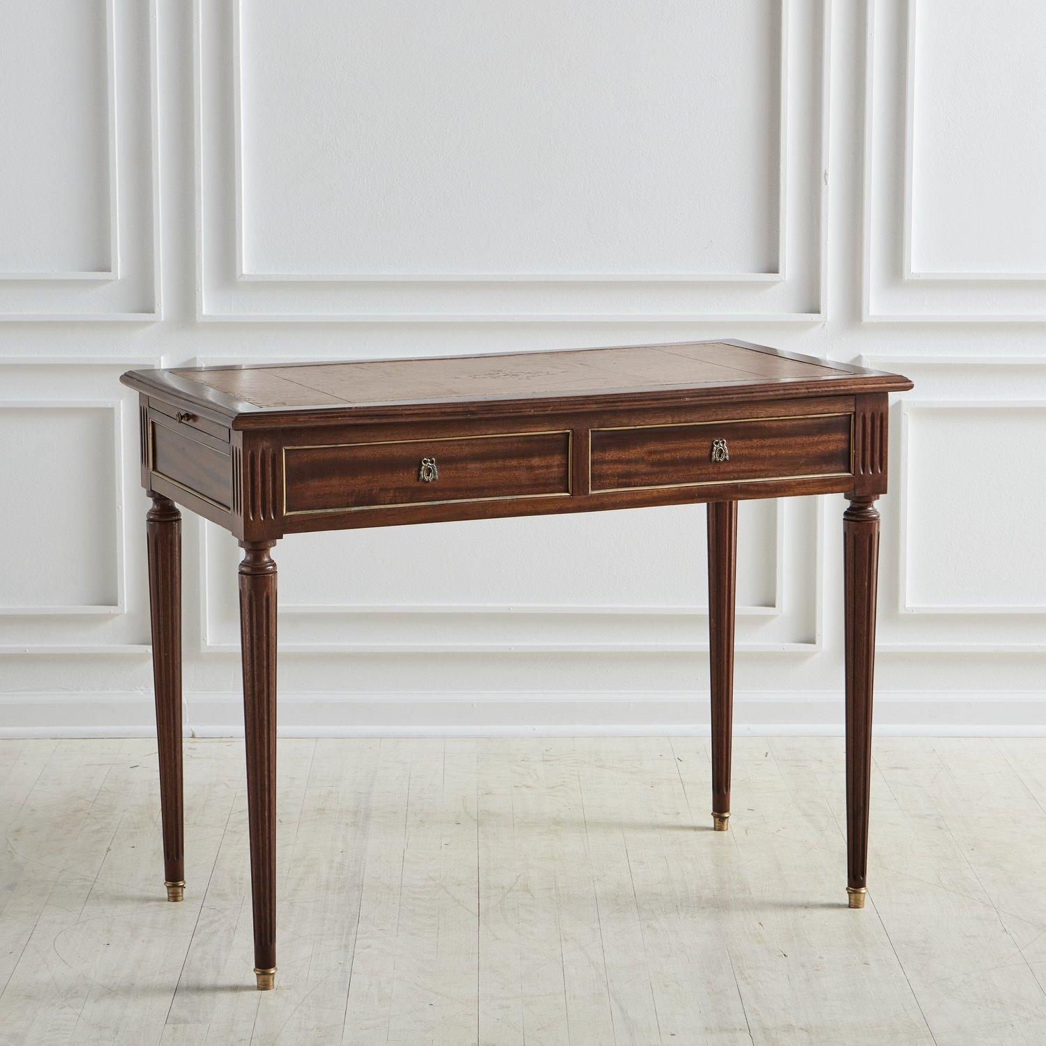A gorgeous Louis XVI style writing desk constructed with mahogany. This desk has a rectangular tabletop with two leaves that pull out for additional surface space. It features an inset cognac leather top with intricate, hand-applied gold leaf