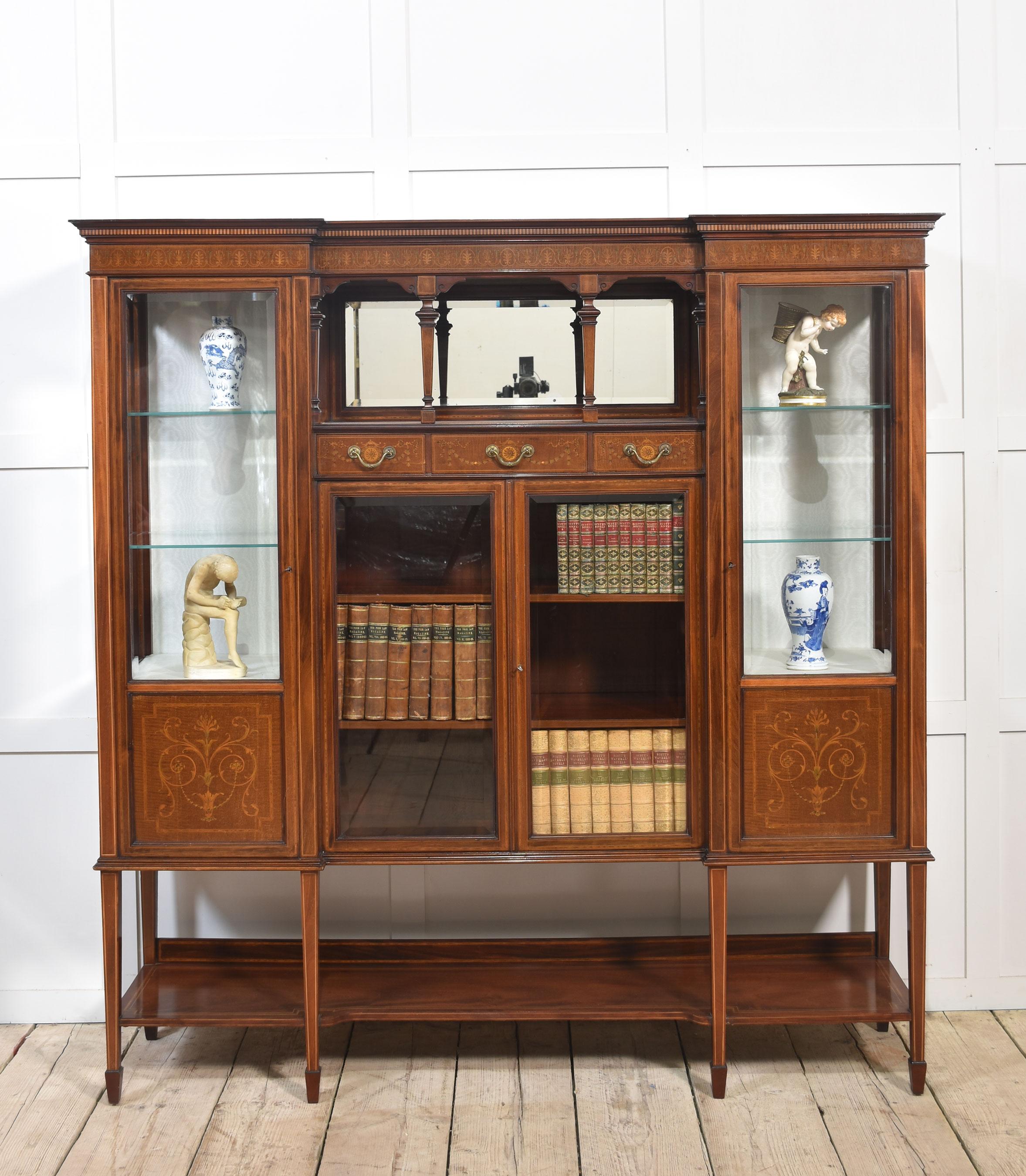 Mahogany inlaid inverted breakfront display cabinet, the moulded top to the central bevelled mirror back arcaded recess above three small drawers and a pair of glazed doors below flanked by a pair of glazed cabinet doors enclosing a lined interior