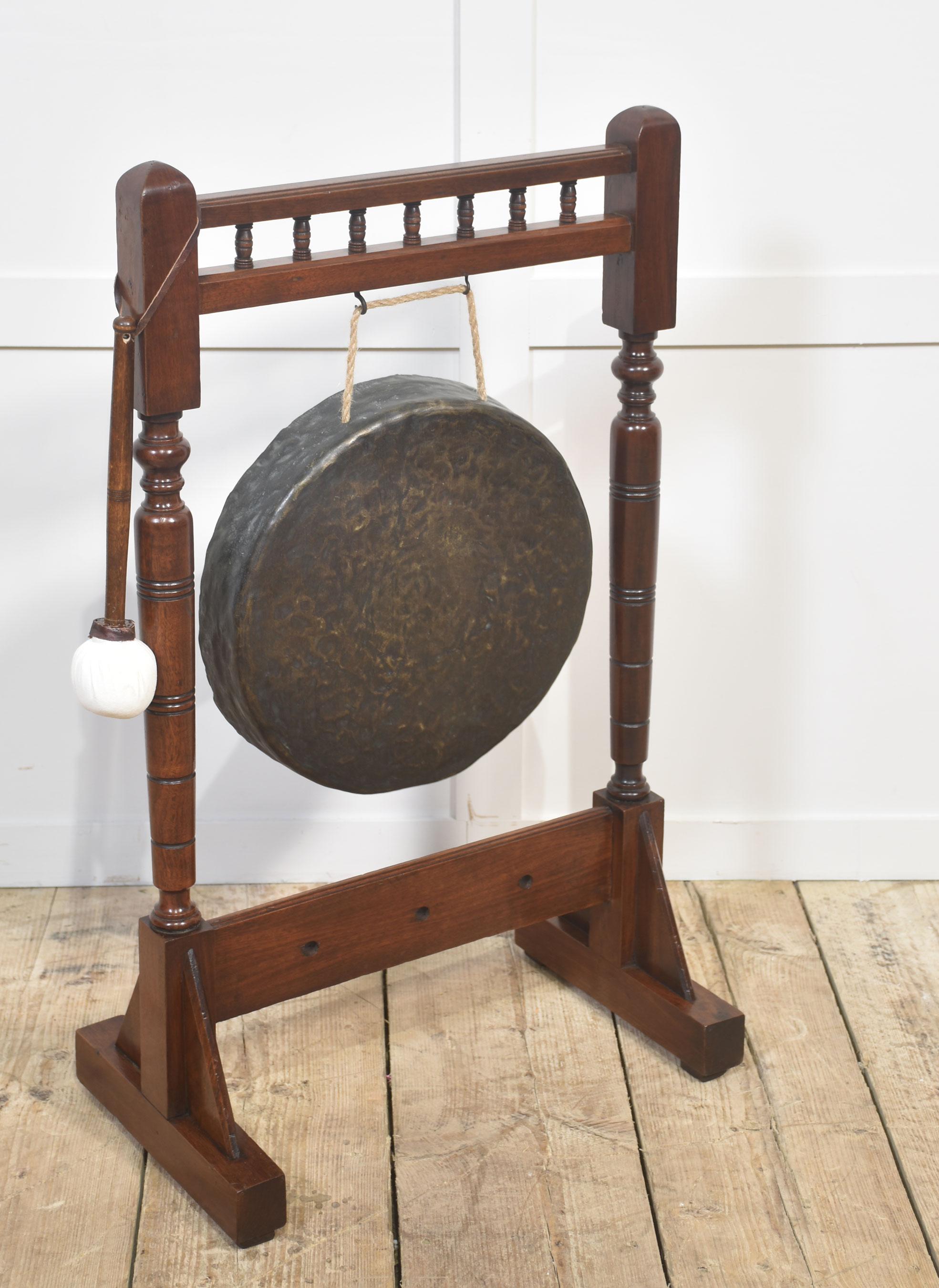 A charming antique dinner gong presented on an elegant, turned mahogany stand. The circular hammered brass gong is suspended from a rope cord and produces a deep, resonant tone when struck with the accompanying mallet. The beautifully crafted wooden