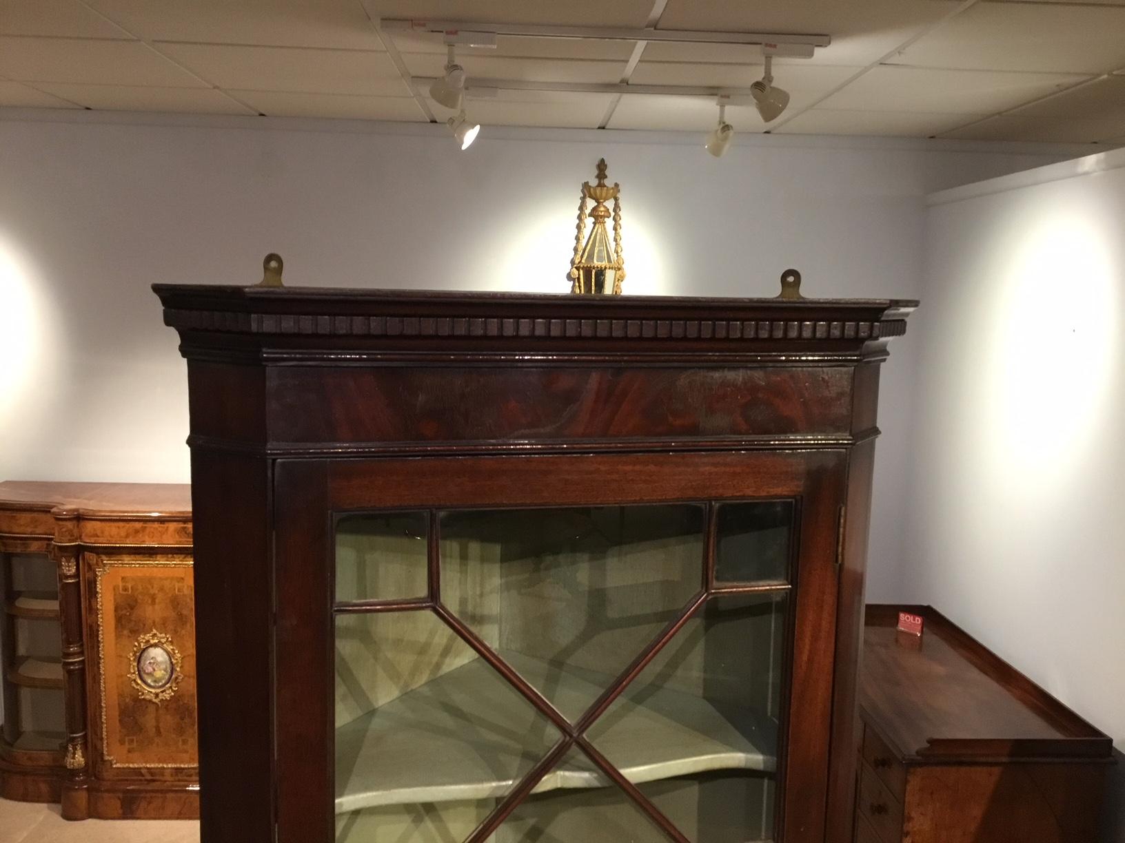 A mahogany George III Period astragal glazed hanging corner cabinet. The dentil moulded cornice above a flame mahogany frieze. Having an astragal glazed door, opening to reveal a painted interior with two shaped shelves, English, circa