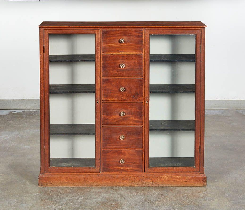 Una libreria indipendente in mogano con cassetti portaoggetti che poggia su uno zoccolo in legno. Due ante integrali vetrate davanti a quattro ripiani per lato e al centro una pila di sei cassetti con pomelli in bronzo in mogano fiammato. Una cassa