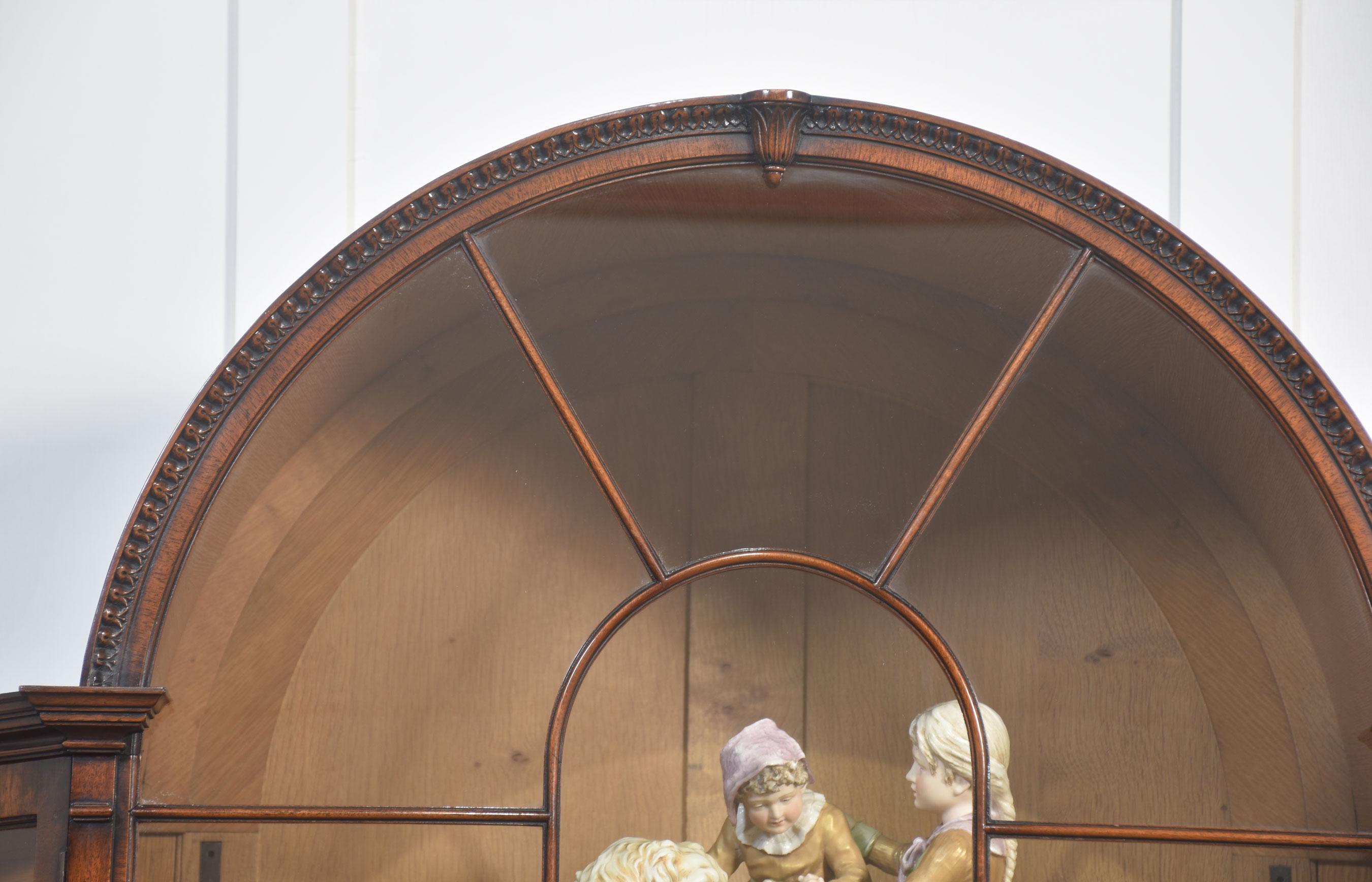 Vitrina de caoba, la parte superior arqueada sobre dos inusuales puertas laterales cóncavas acristaladas, el interior equipado con estantes acristalados ajustables. En la base, tres largos cajones centrales con tiradores de latón en forma de cuello