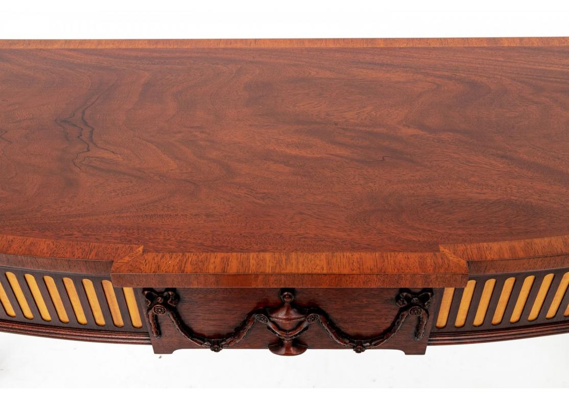A Henkel-Harris Neo-Classical style console table. Mahogany with shaped top over fluted skirt with contrasting stains and central relief carved crest depicting an urn and floral garland. Raised on tapering and fluted legs ending in spade feet. Cloth
