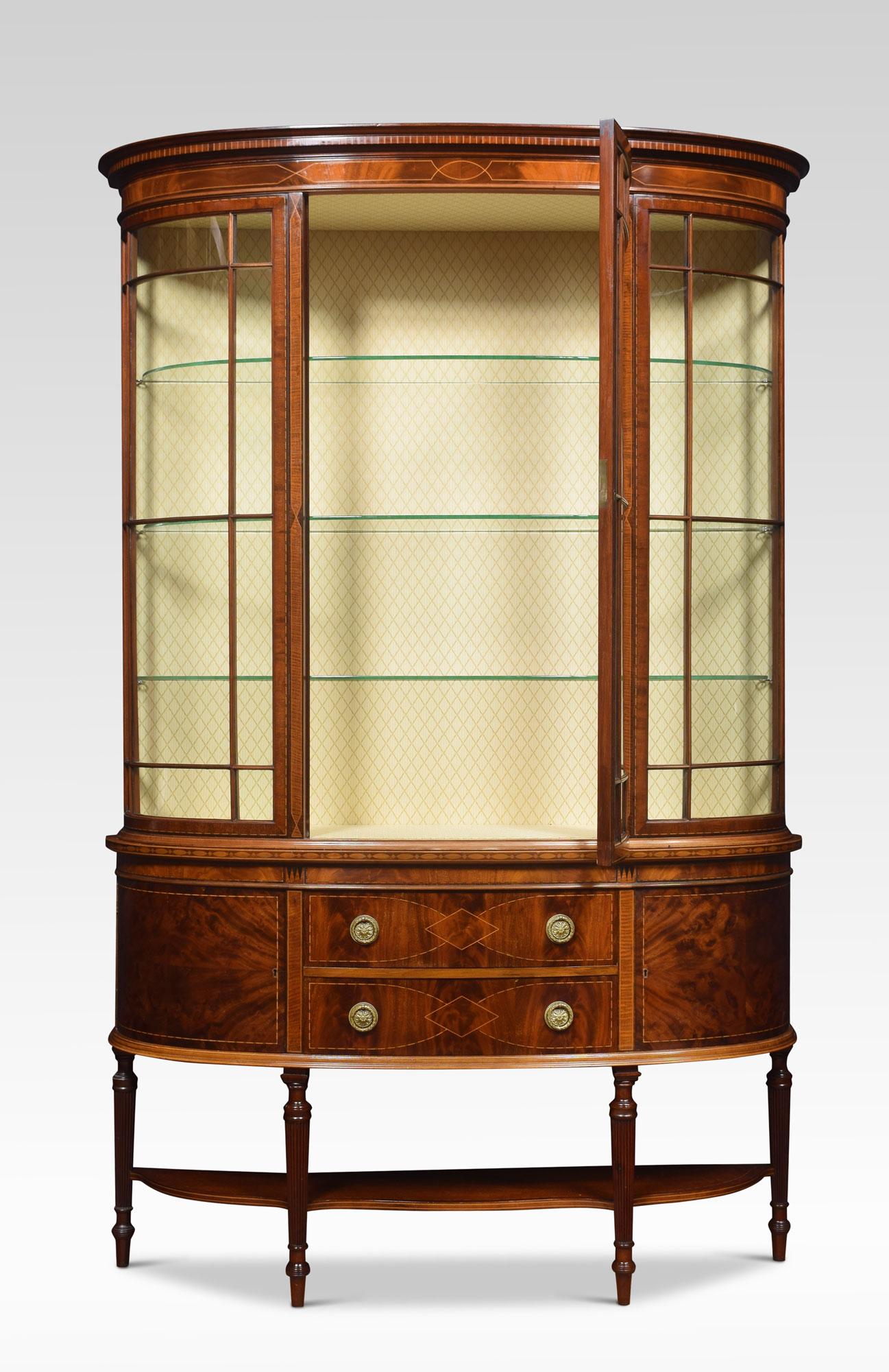 Mahogany bow fronted display cabinet the moulded cornice above large central single door opening to reveal the upholstered interior and three glazed shelves. The base section fitted with two central drawers having tooled brass handles flanked by