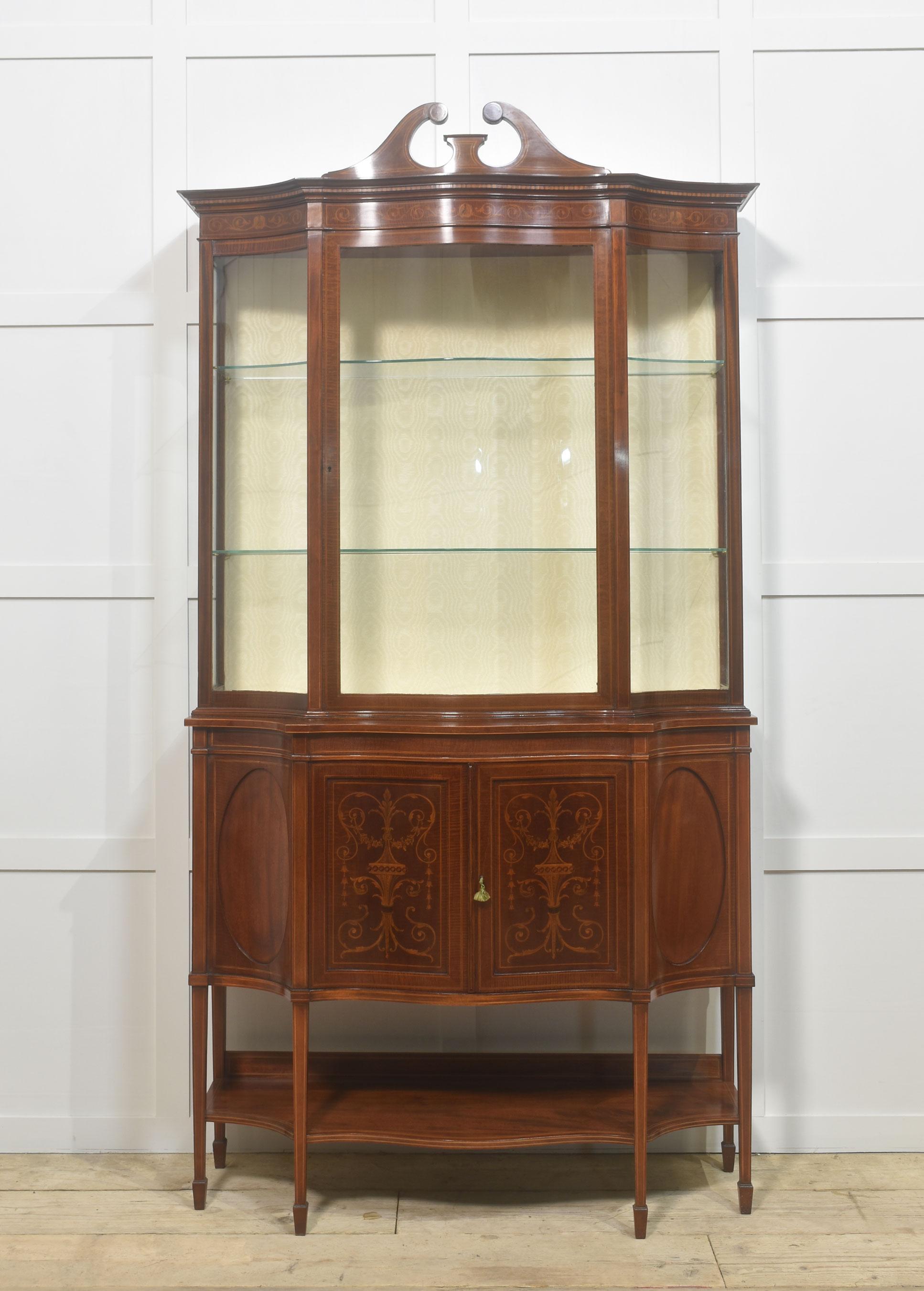 Mahogany inlaid display cabinet the swan neck pediment above a pair of glazed doors opening to reveal upholstered silk interior with two glazed shelves. flanked by serpentine ends. To the base section with a pair of paneled doors with floral inlay