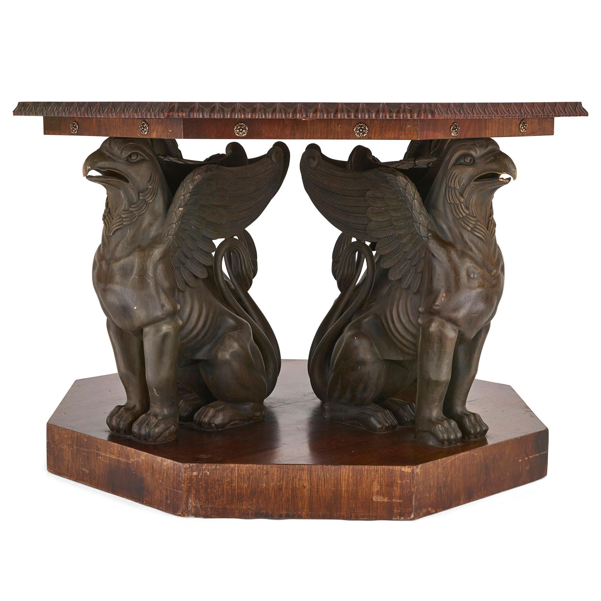 The octagonal top of this table, of solid mahogany, sits above an octagonal frieze, decorated with rosettes, which in turn is supported by four bronzed metal seated griffins. The griffins sit upon a solid mahogany octagonal plinth base. The design