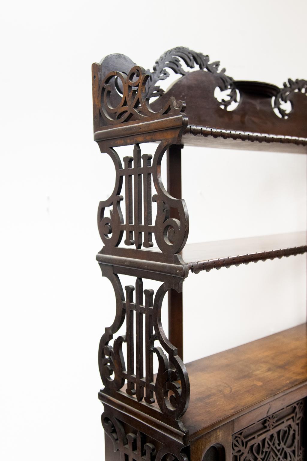 Mahogany open fretwork three-tiered shelf has sides of stylized lyre shape. The two doors with blind fretwork fold down. The middle shelf and top have applied carved moldings. The top crest has carved open work floral arabesques.