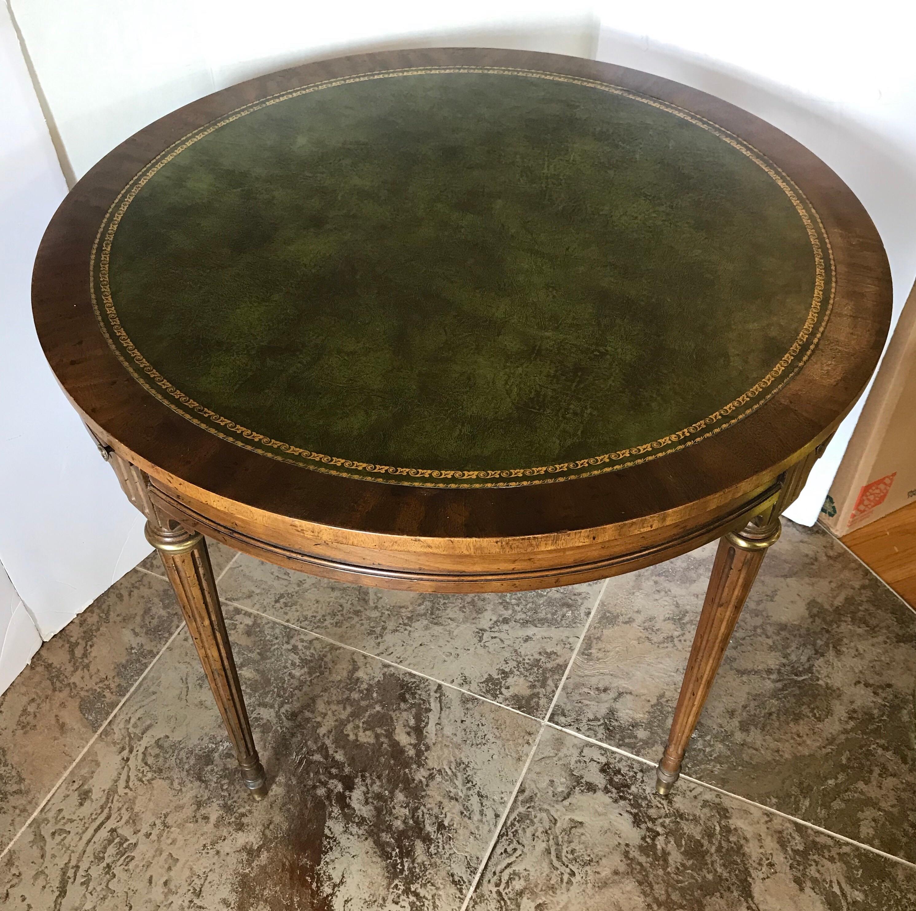 Round mahogany game table has a leather top in dark green with decorative gold border. Above each leg is a pull out drink holder with a brass pull. The table is supported by fluted tapered legs each having a round brass mount.