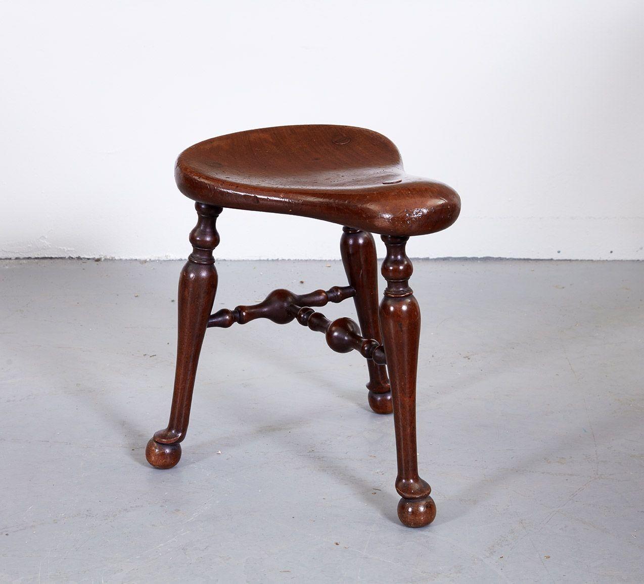 Good late 19th century mahogany saddle stool by James Shoolbred and Co. having shaped seat over turned legs ending in pad feet and joined by bulbous turned T-stretcher, the whole with good rich color and patination, the underside stamped with