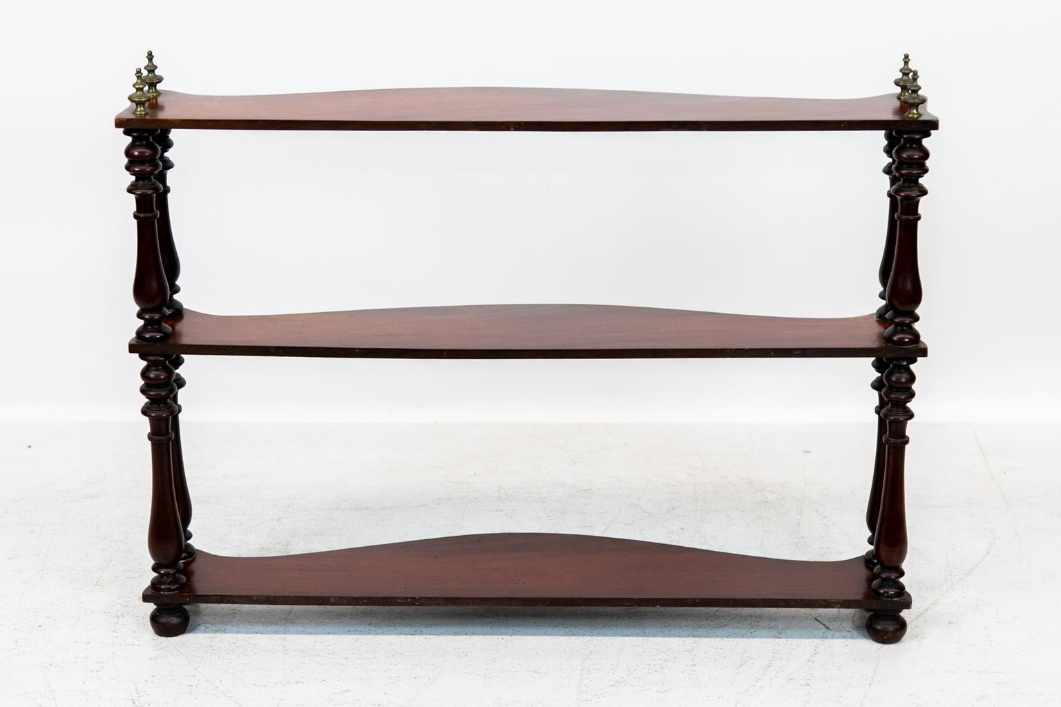 Mahogany serpentine three-tier shelf, has four brass finials and is supported on bun feet. It is designed for hanging or counter top use.
  
