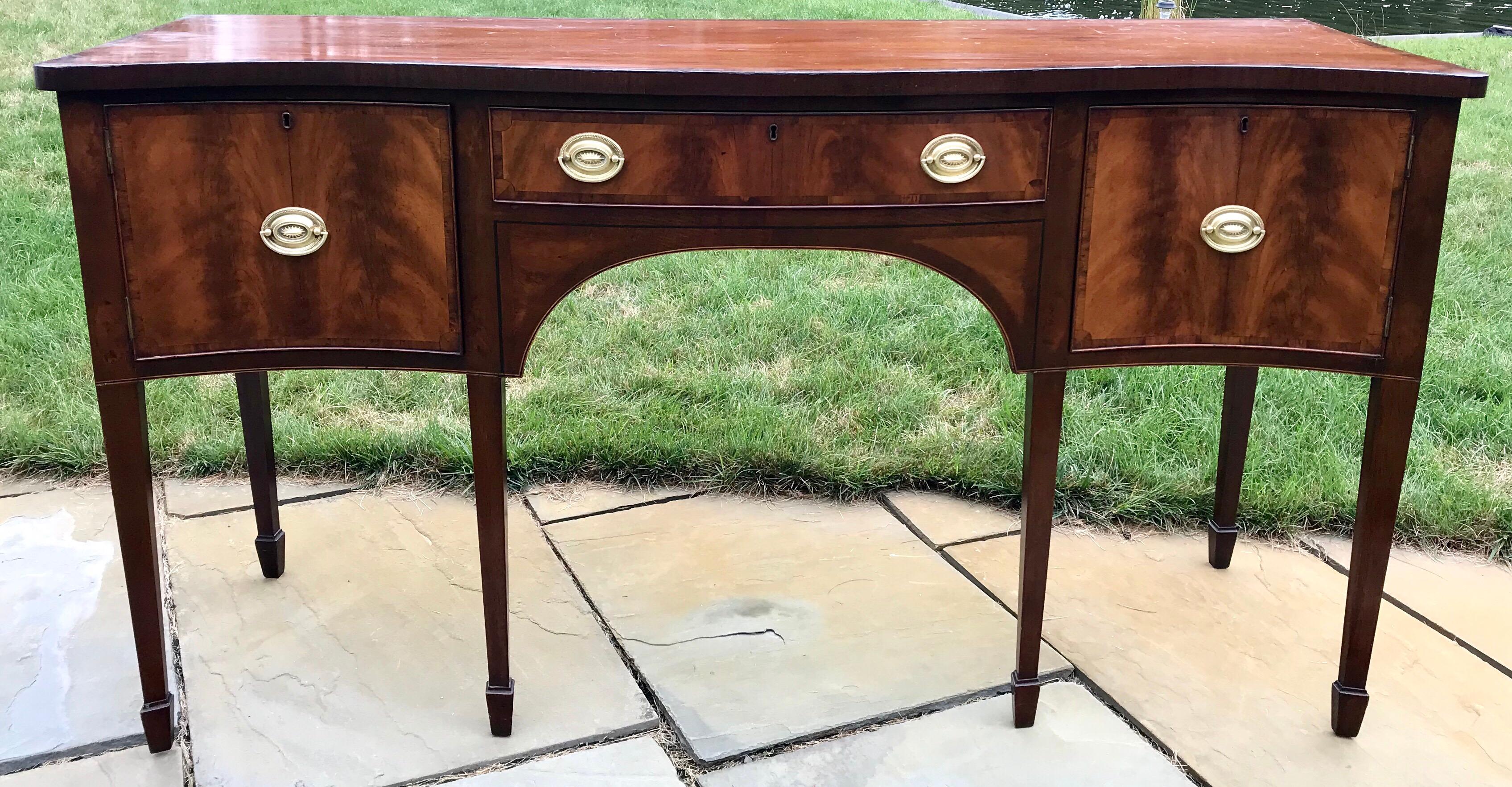 George III period English serpentine front sideboard with beautifully bookmatched crotch grain Mahogany veneered front doors and drawer. The tapered legs have spade feet which is typical for Hepplewhite's design.