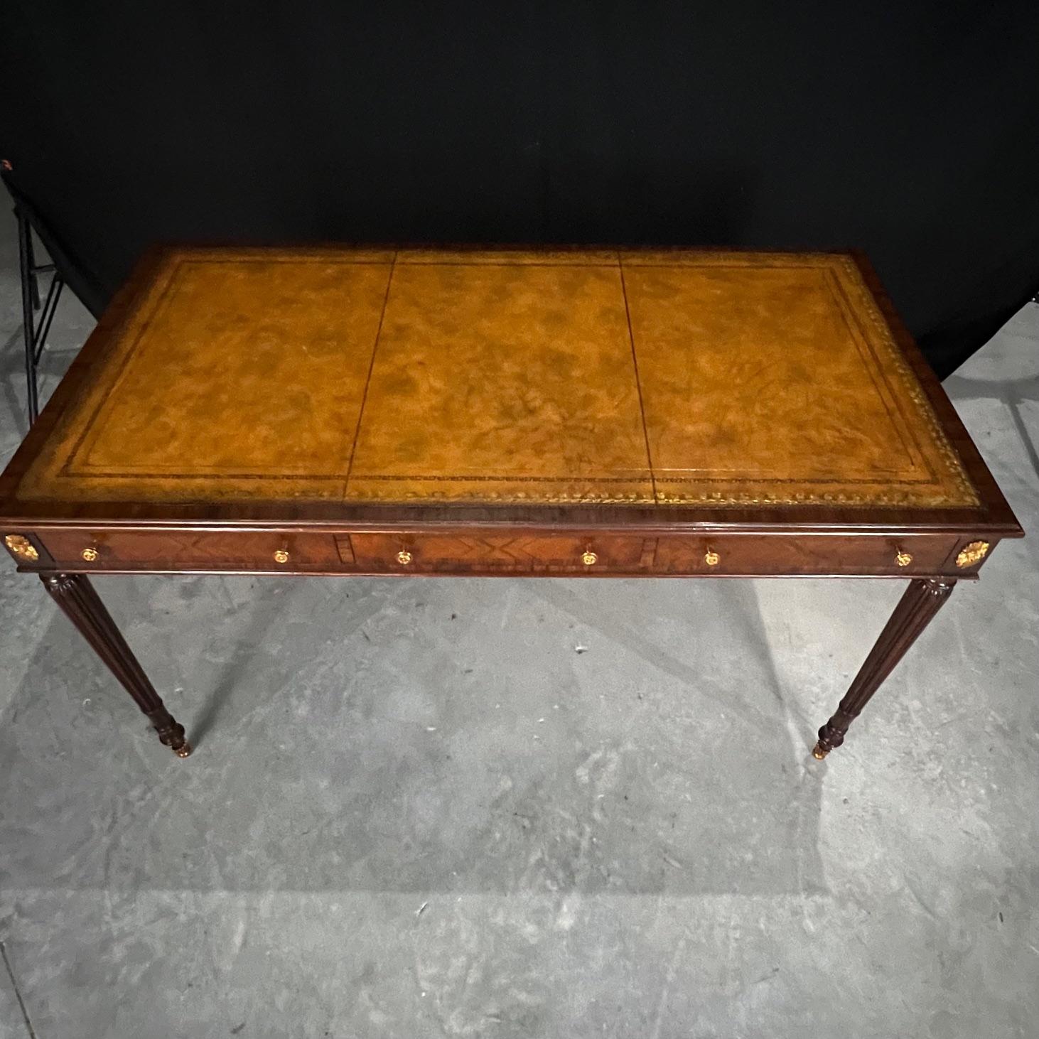 Elegant rectangular mahogany writing desk with three panel embossed tooled leather top above three fitted drawers with intricate brass knobs, supported by four tapered reeded legs with brass ormolu on all sides. A Maitland Smith label in the center