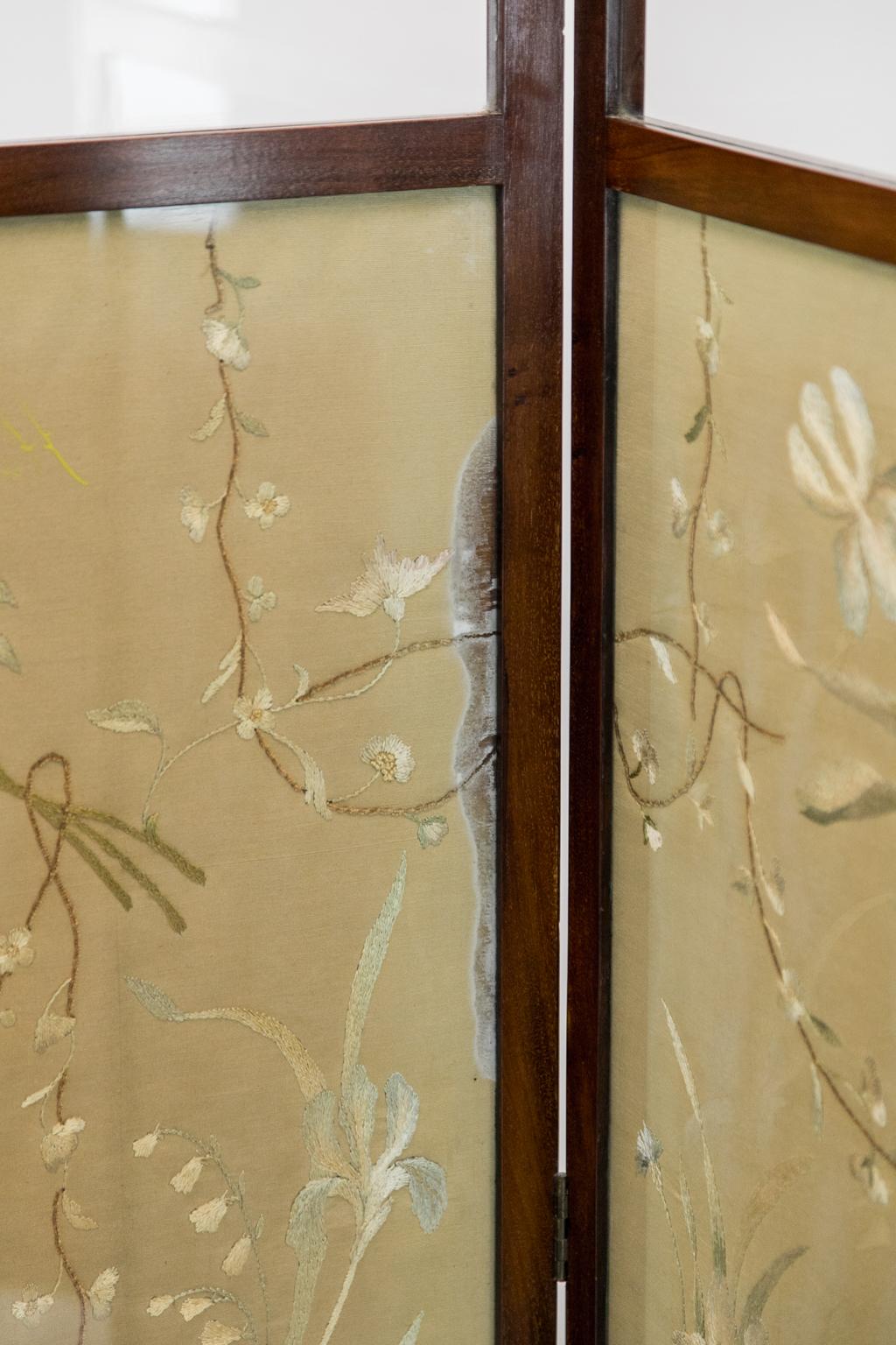 This solid mahogany folding screen has the original clear, wavy glass in the top panels. The silk needlework depicts groupings of floral vines and bundles of daisies, lily of the valley, and other flowers. The left-hand panel has a 11 1/2