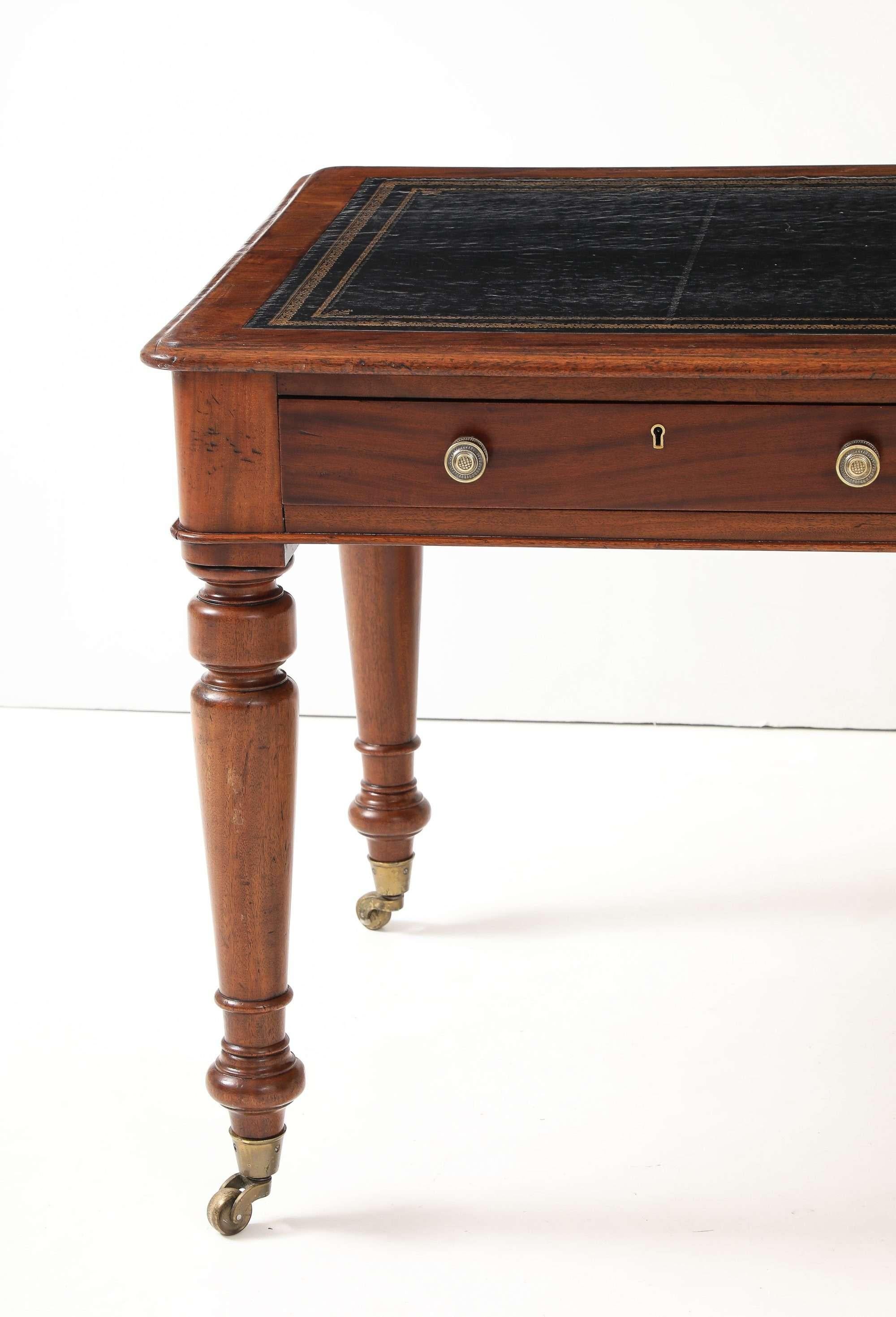 A handsome mid-19th-century English writing table featuring a rectangular mahogany frame inset with a black, gilt-tooled leather writing surface. Two locking frieze drawers retain their original brass knob pulls, while ring-turned, tapering legs