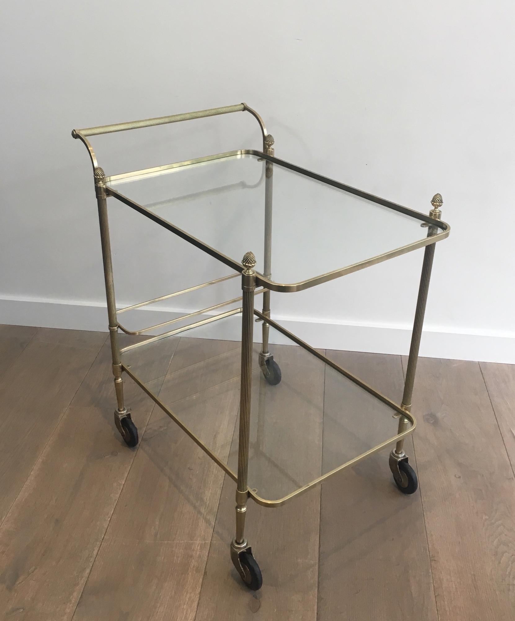 This neoclassical bar cart is made of brass with 4 finials and 2 clear glass shelves. This drinks trolley has a bottles holder on bottom shelf. This is a work by famous French designer Maison Bagués, circa 1940.