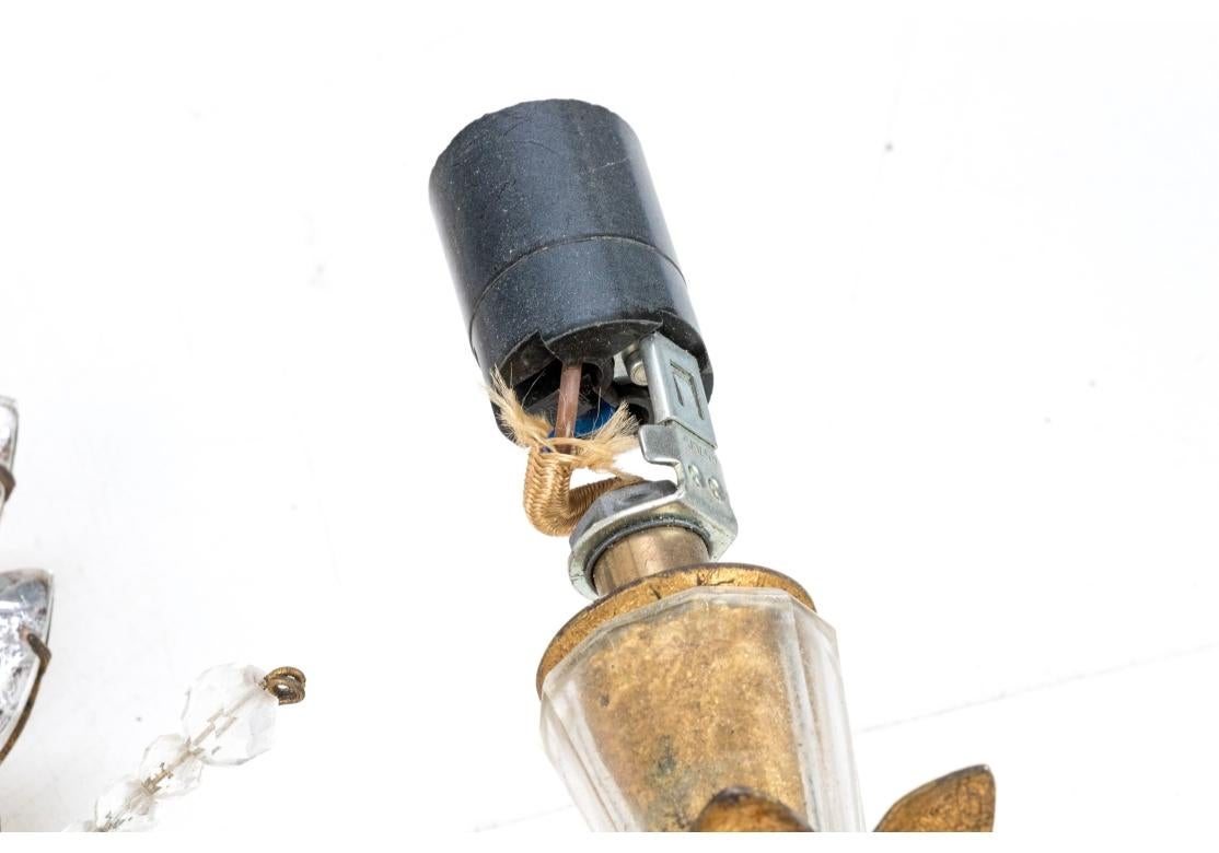 Pareja de apliques de pared de dos luces de metal dorado con una flor central de cristal facetado, una rama de hojas debajo y encima con incrustaciones de hojas de cristal moldeado y cuatro puntas emisoras de cuentas facetadas. Los portavelas tienen