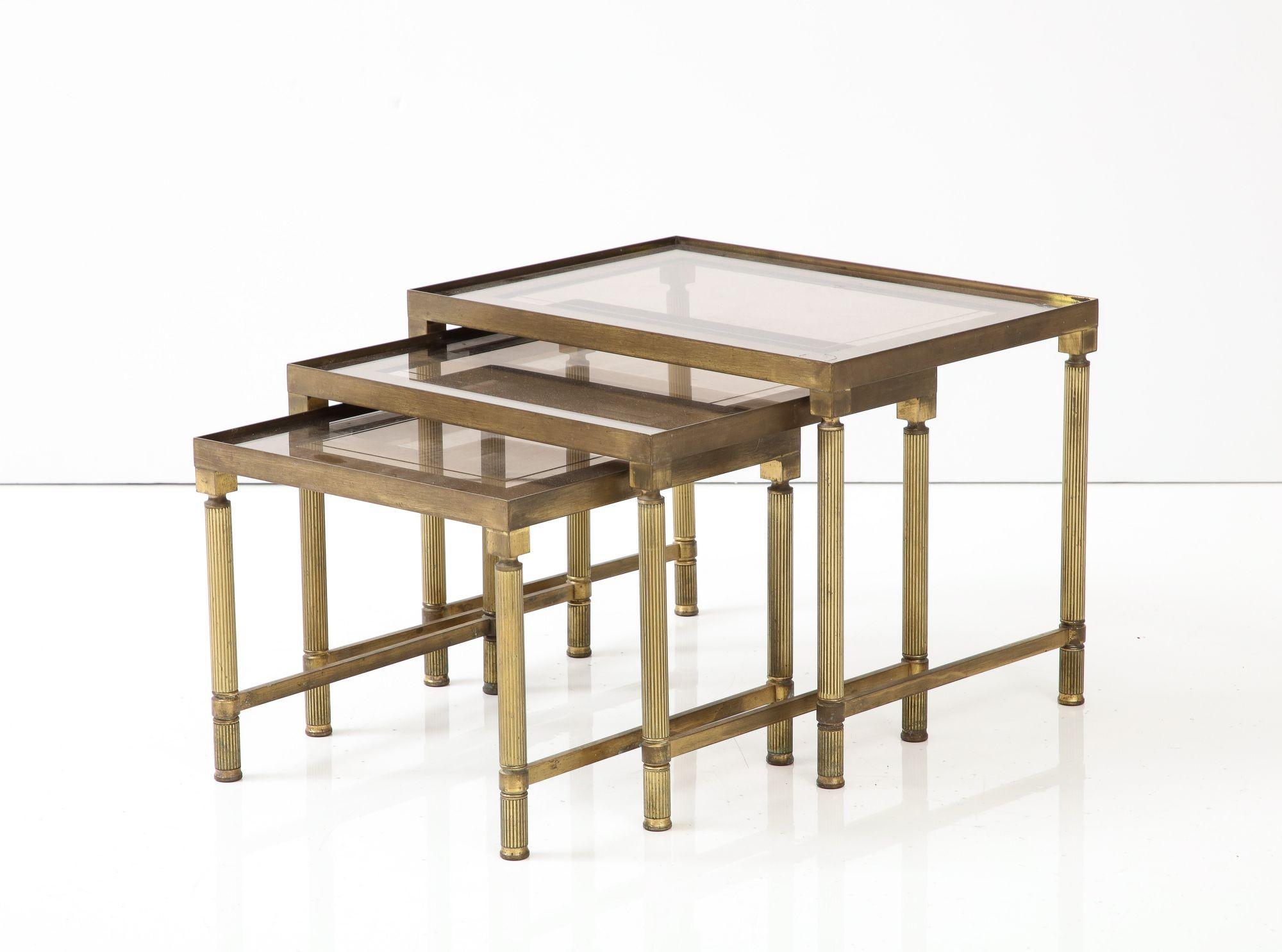 This trio of mid-20th-century French Maison Bagues nesting tables epitomizes timeless sophistication and exquisite craftsmanship. Supported by timeworn brass legs and stretchers, these tables exude opulence. Their smoked glass tops, adorned with a