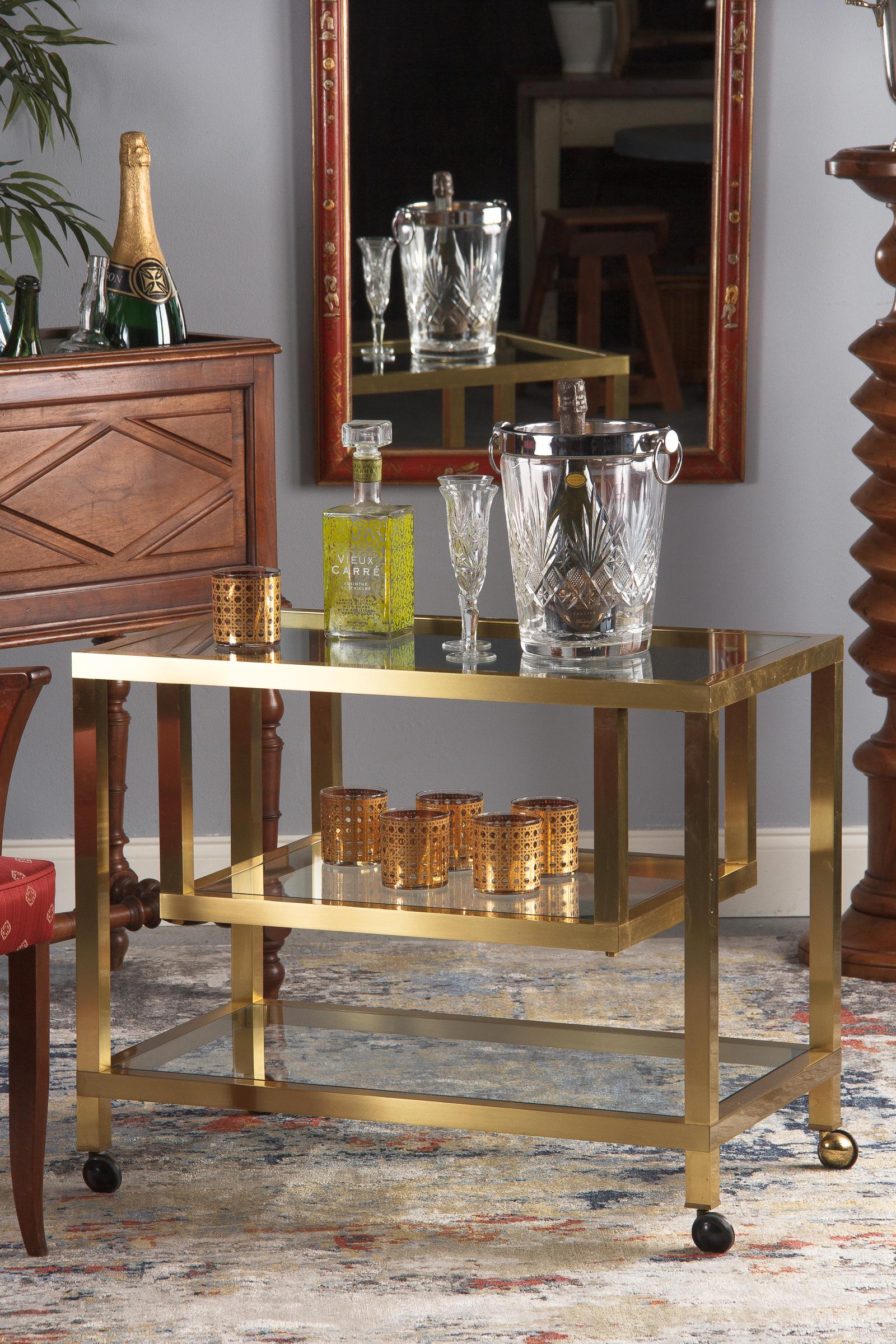 A stylish three-tiered vintage brass and glass bar cart attributed to Maison Jansen, circa 1970. The unusual brushed brass frame has a rectangular shape with a top and bottom tier, and a smaller middle tier that is suspended from the top. All three